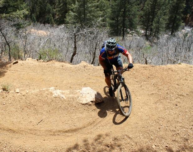 Rocky Mountain Instinct 999 MSL: A mountain biker navigating a curved dirt trail surrounded by trees. The cyclist is wearing a helmet and biking gear, demonstrating skill as they maneuver around a rock on the path. The landscape features varying shades of brown and green, typical of a forested area on a sunny day.