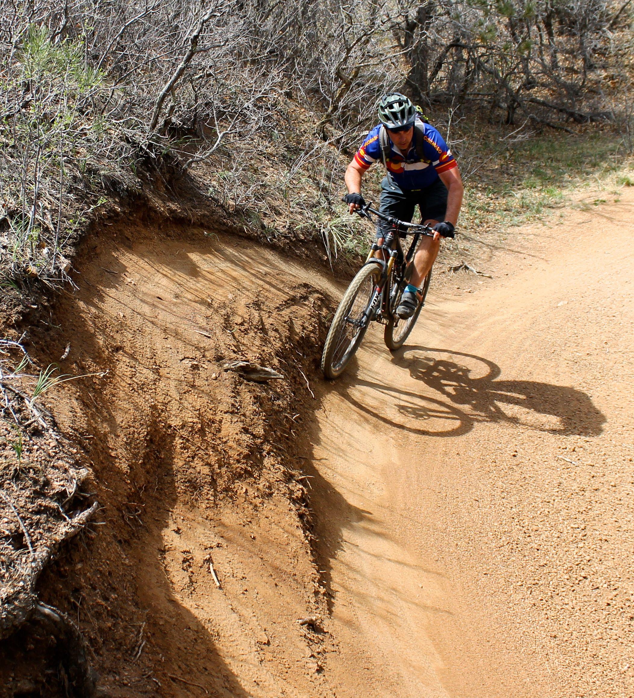 A mountain biker skillfully navigates a dirt trail, leaning into a turn on a curved section of the path. The surrounding terrain features sparse vegetation and dry, sandy soil. Stratton Open Space / The Chutes mountain bike trail.