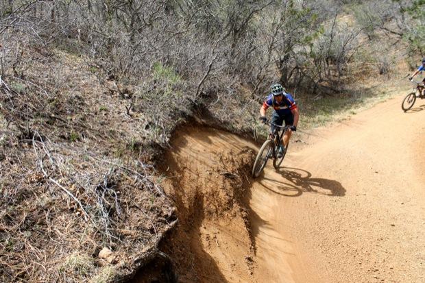 Rocky Mountain Instinct 999 MSL: A mountain biker navigates a dirt trail with a sharp turn, surrounded by sparse vegetation and rocky terrain. Another cyclist can be seen in the background.