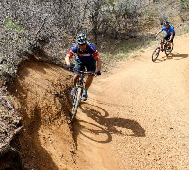 Rocky Mountain Instinct 999 MSL: Two mountain bikers navigate a curved dirt trail surrounded by sparse vegetation. One rider is leaning into a turn, showcasing an athletic position, while the other follows closely behind on a parallel path. The sun highlights the texture of the terrain and the riders' vibrant cycling attire.