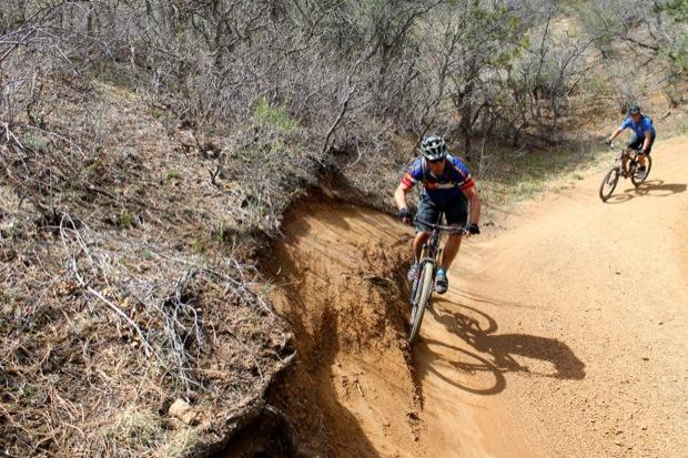 Rocky Mountain Instinct 999 MSL: Two mountain bikers navigate a dirt trail in a forested area. One rider leans into a turn while descending, showcasing their skill on the uneven terrain. The second rider follows closely behind on the winding path, surrounded by sparse vegetation and earthy tones.