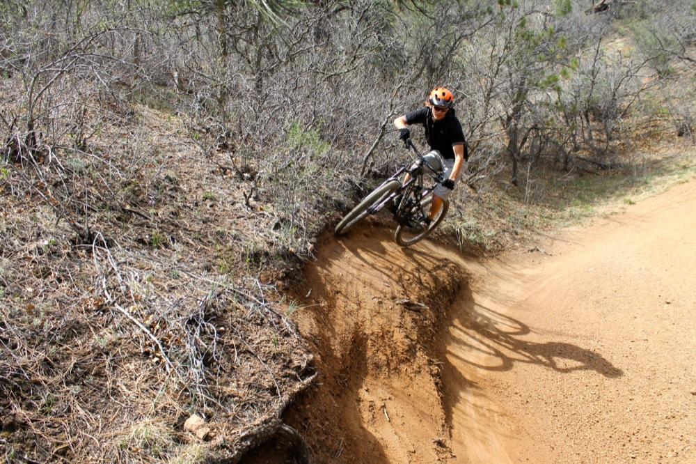A mountain biker in an orange helmet rides aggressively along a dirt trail, leaning into a sharp turn. Surrounding the trail is dry brush and sparse vegetation, with the bike's wheels slightly off the ground, showcasing the rider's skill and balance. Stratton Open Space / The Chutes mountain bike trail.