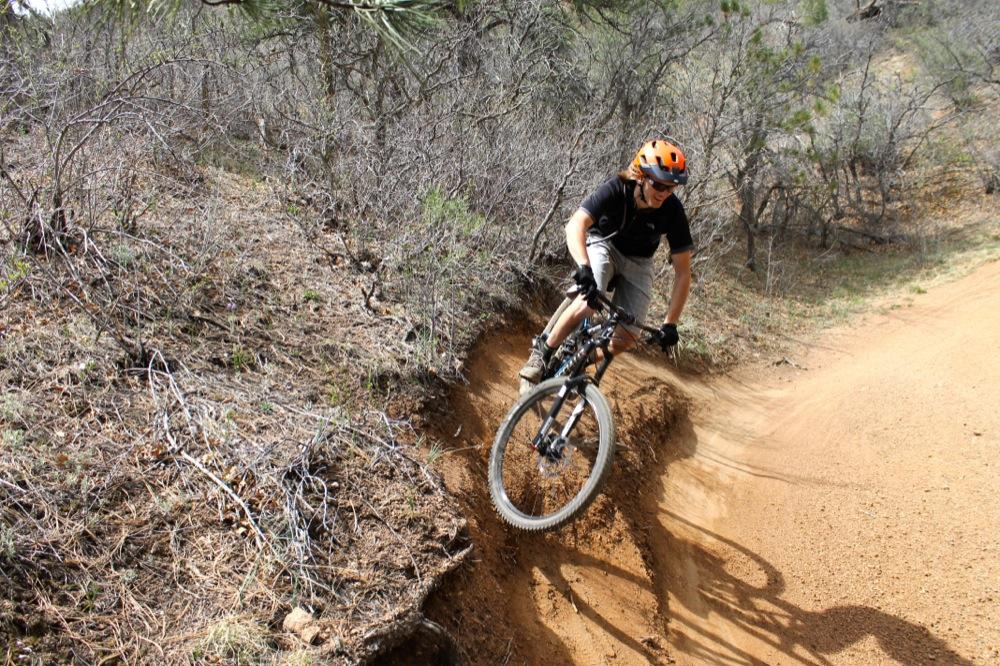 A mountain biker launching off a dirt trail, navigating a curve with one tire off the ground. The background features dry vegetation and sparse trees, suggesting a rugged outdoor setting. The biker is wearing a helmet and protective gear. Stratton Open Space / The Chutes mountain bike trail.