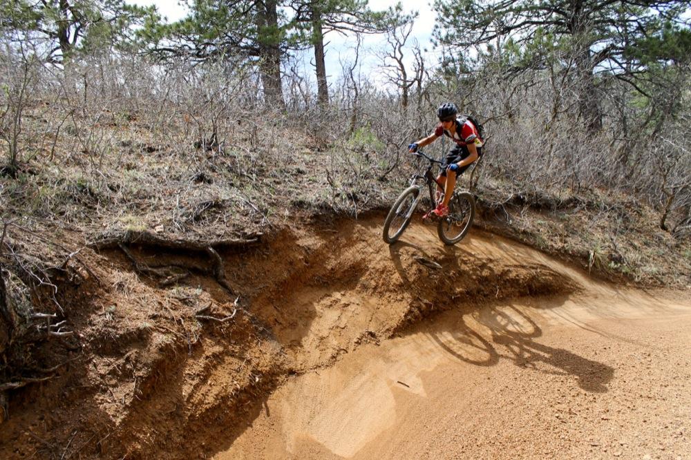 A mountain biker in action, jumping off a dirt jump on a biking trail surrounded by sparse trees and shrubs. The biker is wearing a helmet and protective gear, capturing a dynamic moment against a backdrop of earthy tones and uneven terrain. Stratton Open Space / The Chutes mountain bike trail.