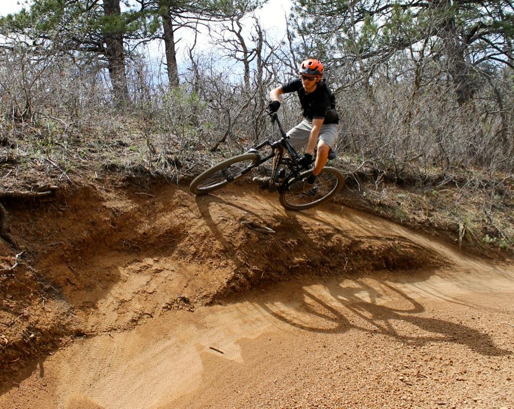 A mountain biker performing a sharp turn on a dirt trail, with a backdrop of sparse trees and brush, showcasing dynamic movement and skill. Stratton Open Space / The Chutes mountain bike trail.