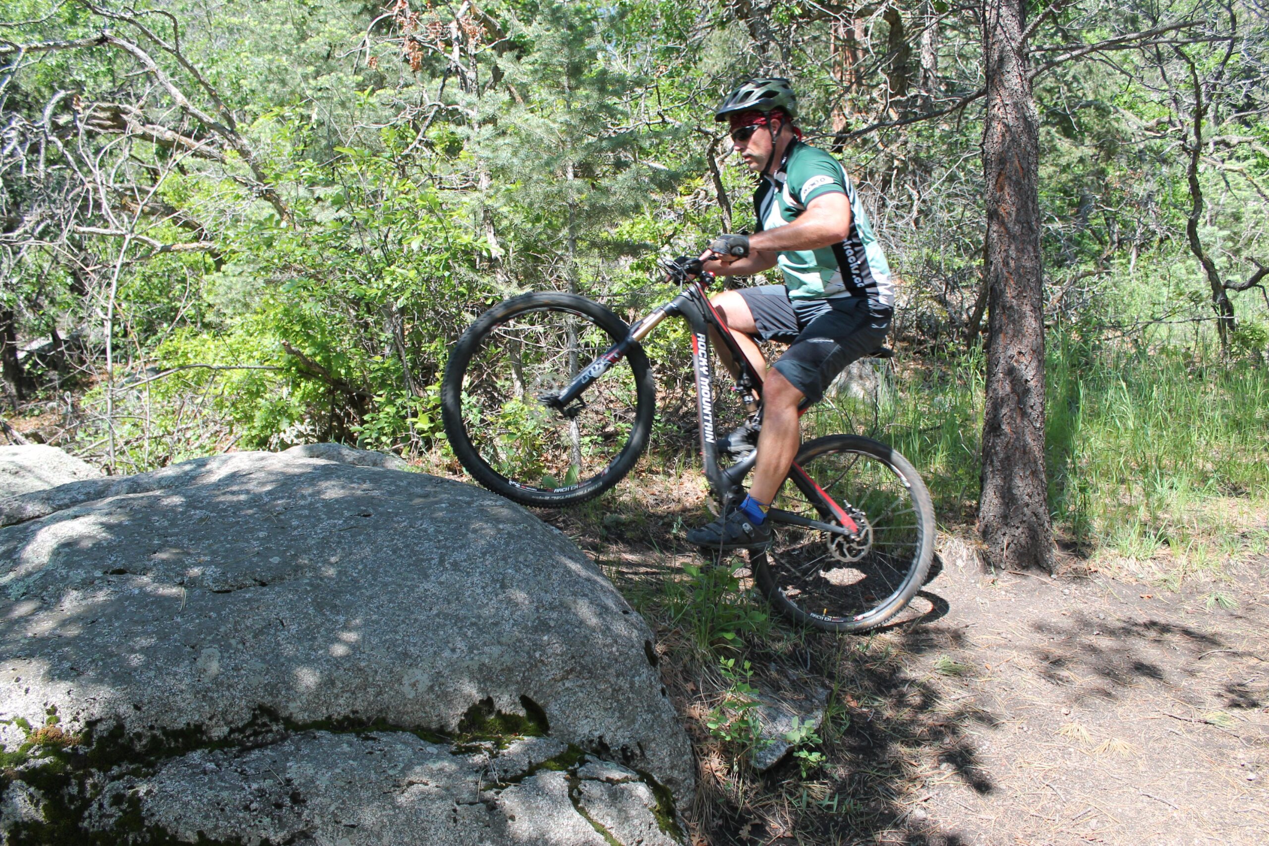 Rocky Mountain Instinct 999 MSL: A mountain biker navigating over a large rock on a forest trail, surrounded by greenery and trees, with a clear blue sky visible above. The biker is wearing a helmet, sunglasses, and a cycling jersey while leaning forward to maintain balance.