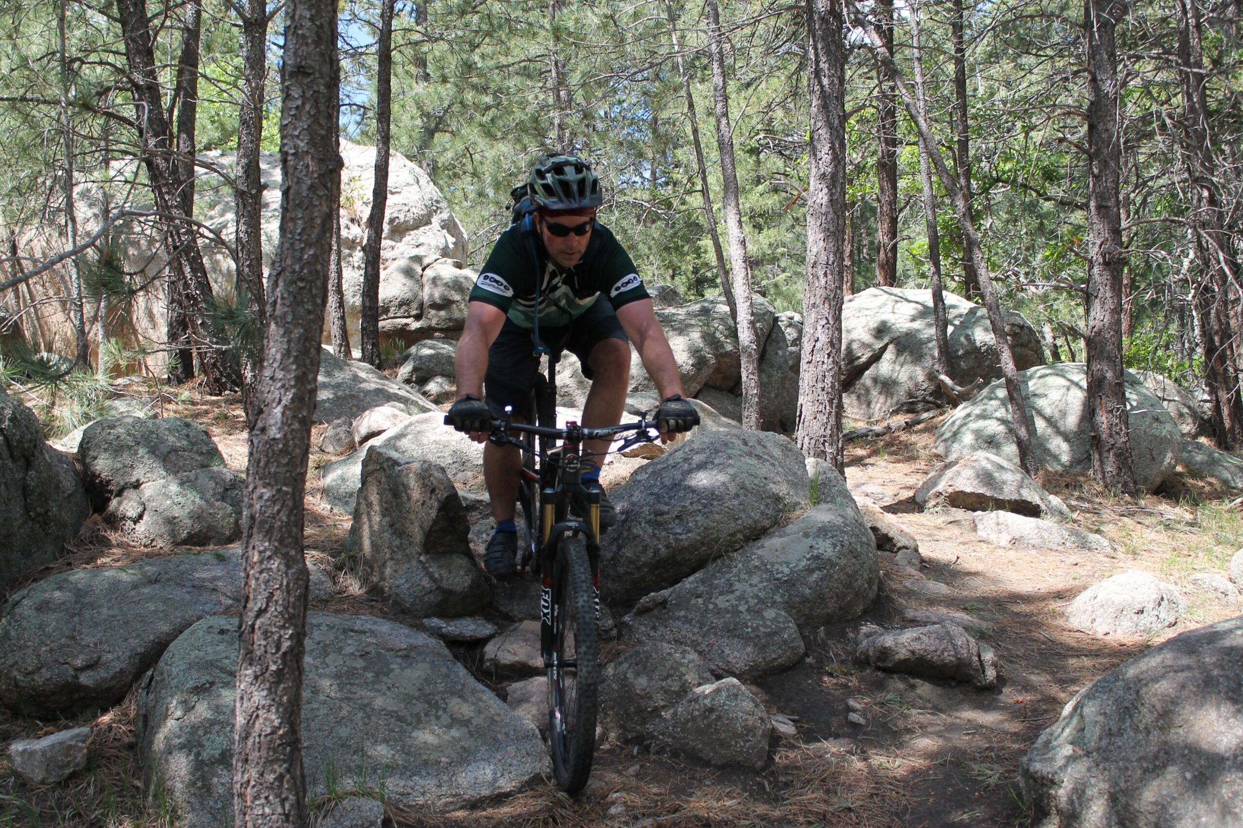 Rocky Mountain Instinct 999 MSL: Mountain biker navigating a rocky trail in a forest, surrounded by trees and boulders on a sunny day.