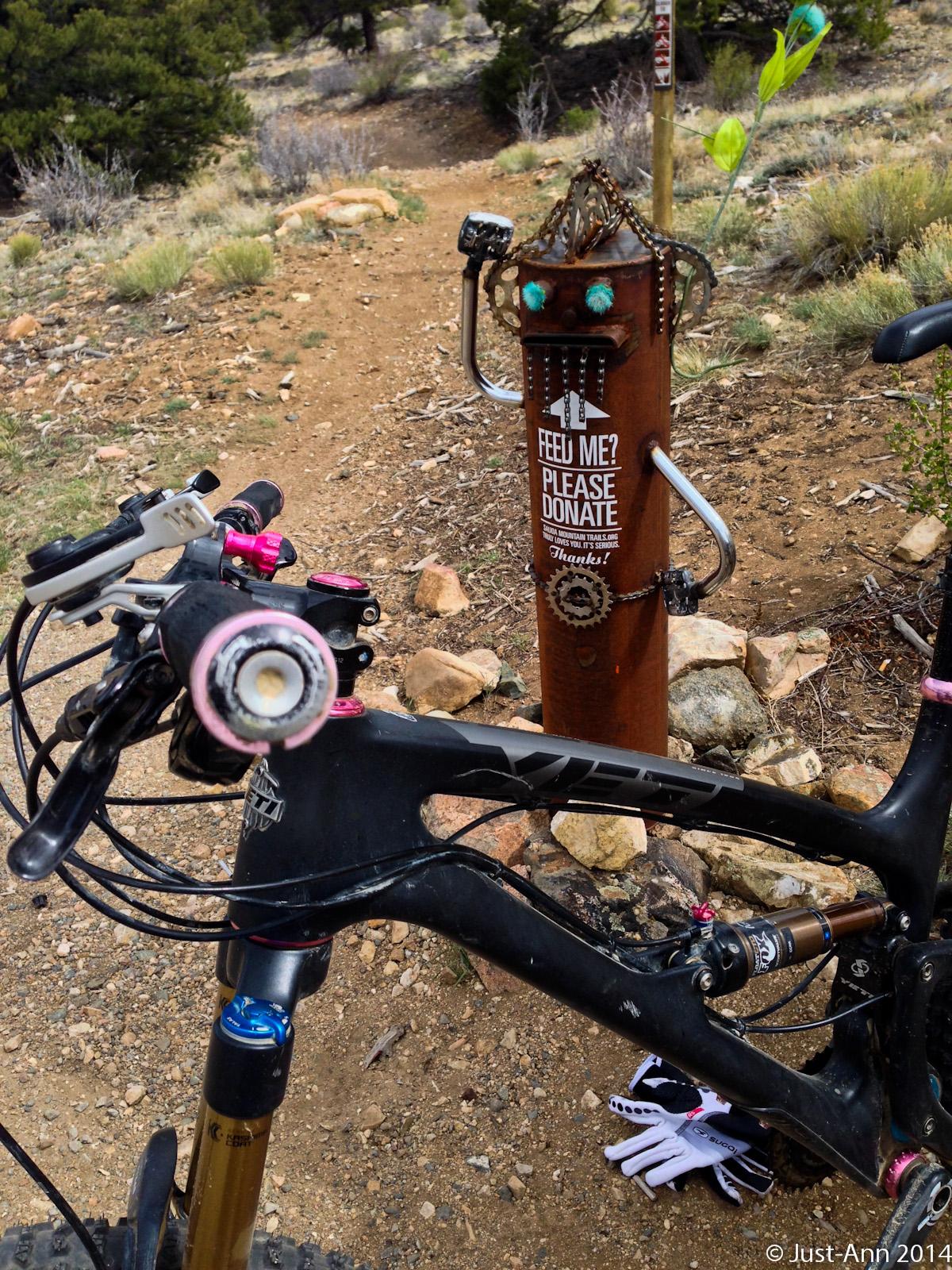 A mountain bike parked near a quirky, artistic donation post along a dirt trail, decorated with whimsical features and a sign that reads "Feel Me? Please Donate. Thanks!" The background features rocky terrain and sparse vegetation, typical of a hiking or biking path. Methodist Mountain mountain bike trail.