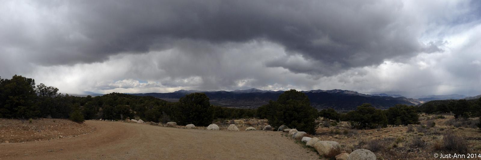 A panoramic view of a landscape featuring dark, stormy clouds above rolling hills and a dirt path lined with rocks. The foreground includes sparse vegetation, while the distant mountains are partially obscured by clouds, creating a dramatic atmosphere. Methodist Mountain mountain bike trail.
