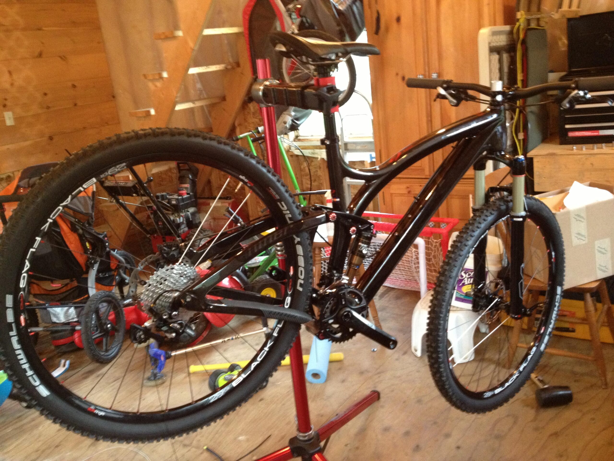 Niner Jet 9 RDO: A black mountain bike is mounted on a bicycle repair stand in a brightly lit wooden workshop. The bike has knobby tires and a visible gear system, with various tools and equipment in the background among boxes and storage items.