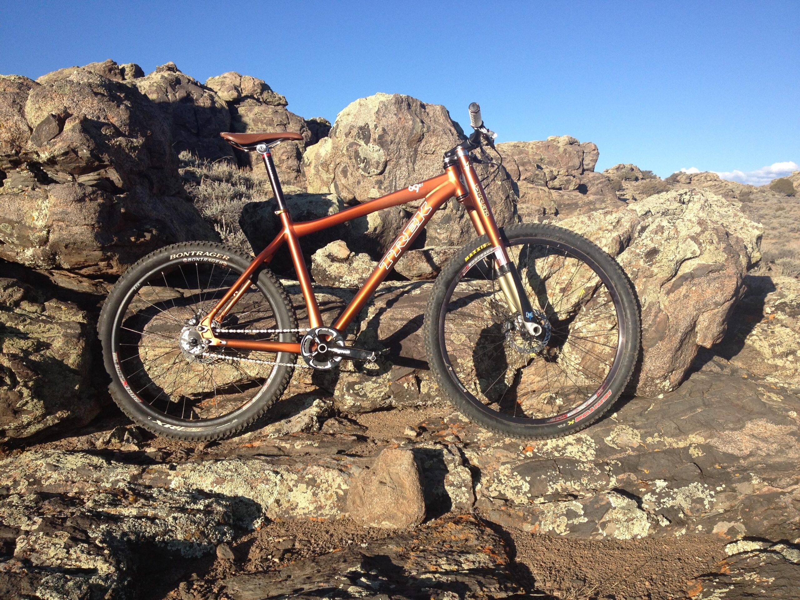 Trek 69er: An orange mountain bike leaning against rocky terrain under a clear blue sky. The bike features thick tires designed for off-road riding, and the surrounding landscape includes rugged rocks and sparse vegetation.