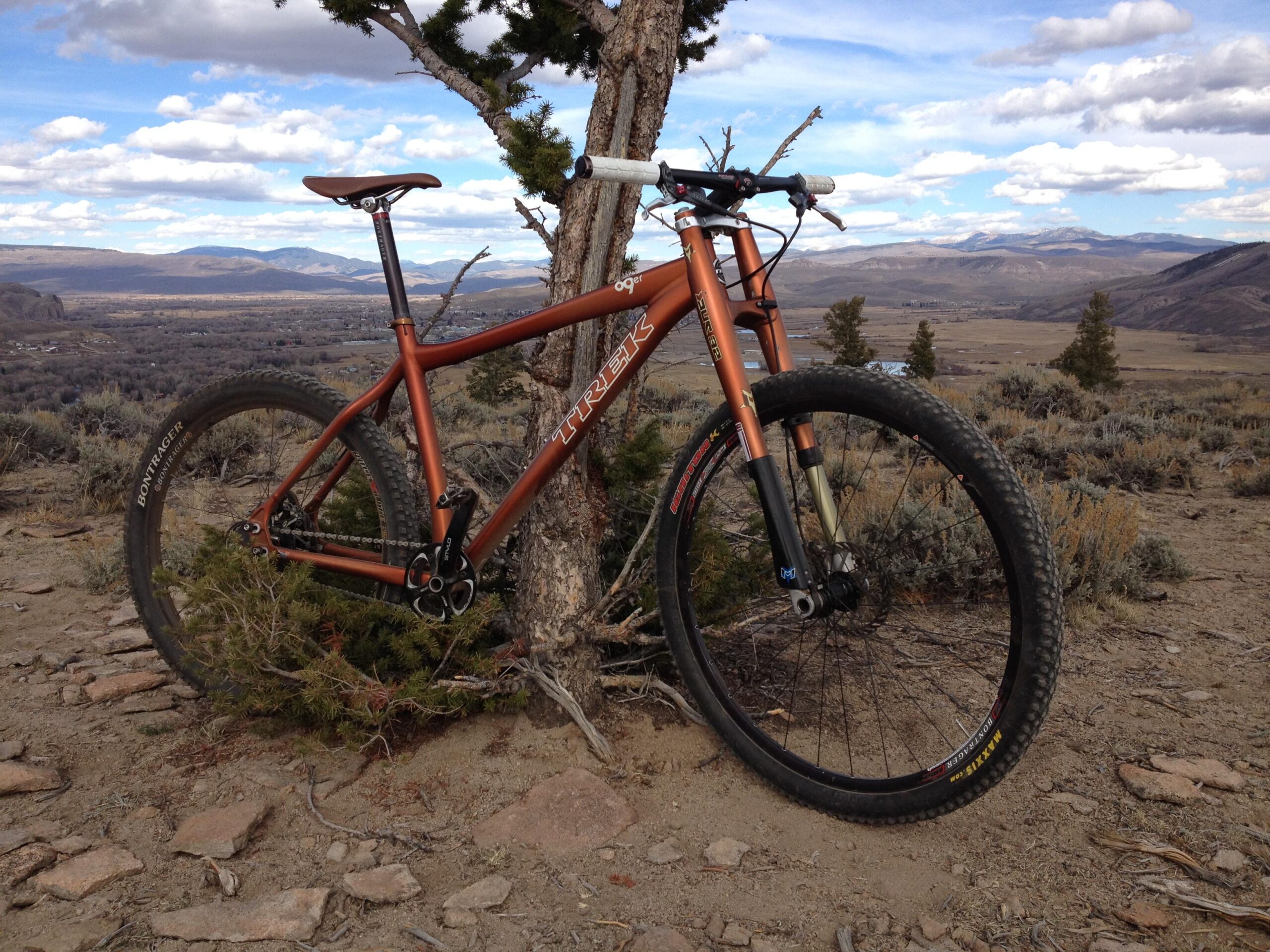 Trek 69er: A mountain bike leaning against a tree on a rocky trail, with a scenic view of mountains and valleys in the background under a partly cloudy sky. The bike has an orange frame and thick tires, surrounded by sparse vegetation and dirt.