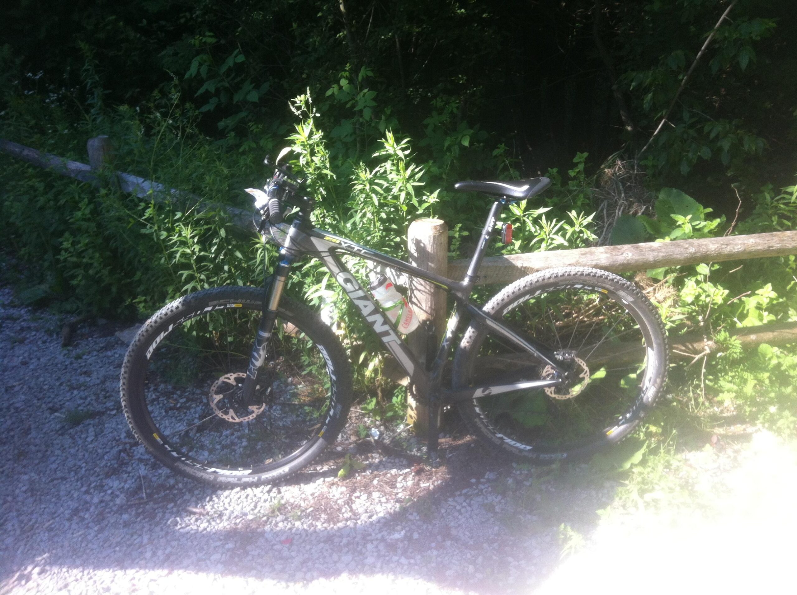 Giant XTC Composite 29er 1: A mountain bike resting against a wooden fence, surrounded by green foliage and gravel terrain. The bike has a black frame with bold white lettering and knobby tires, indicating it's designed for off-road use. Sunlight filters through the trees, casting a gentle glow on the scene.
