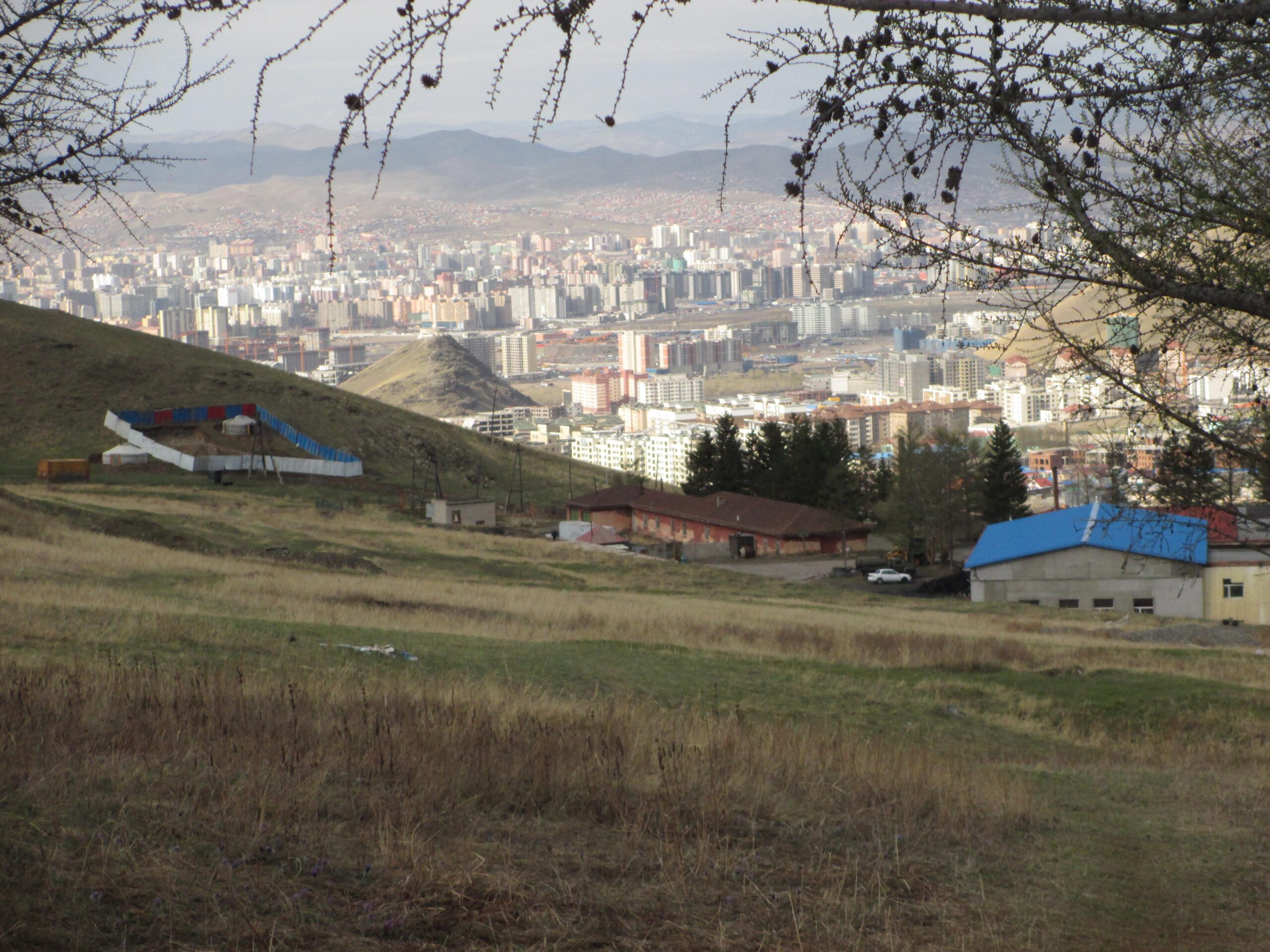 A panoramic view of a city from a hillside, showcasing a blend of urban buildings and natural landscapes. In the foreground, there are grassy slopes and scattered structures, while the background features a sprawling cityscape with numerous high-rise buildings and distant mountains under a cloudy sky. Zaisan Chutes mountain bike trail.
