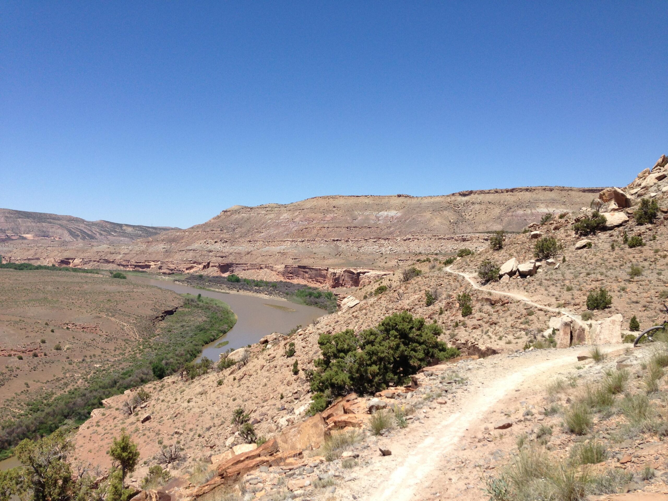A winding dirt trail leads through a rugged, arid landscape featuring brown and green hues, with a winding river visible below. The scene is set under a clear blue sky, with rocky hills in the background. Mary's Loop / Horsethief Bench mountain bike trail.