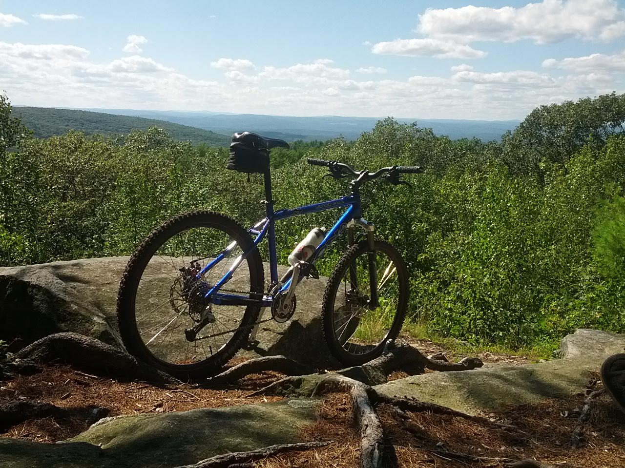 A blue mountain bike rests on a large rock ledge, surrounded by lush greenery. In the background, rolling hills extend towards the horizon under a partly cloudy sky, creating a scenic outdoor landscape. Wendell State Forest mountain bike trail.