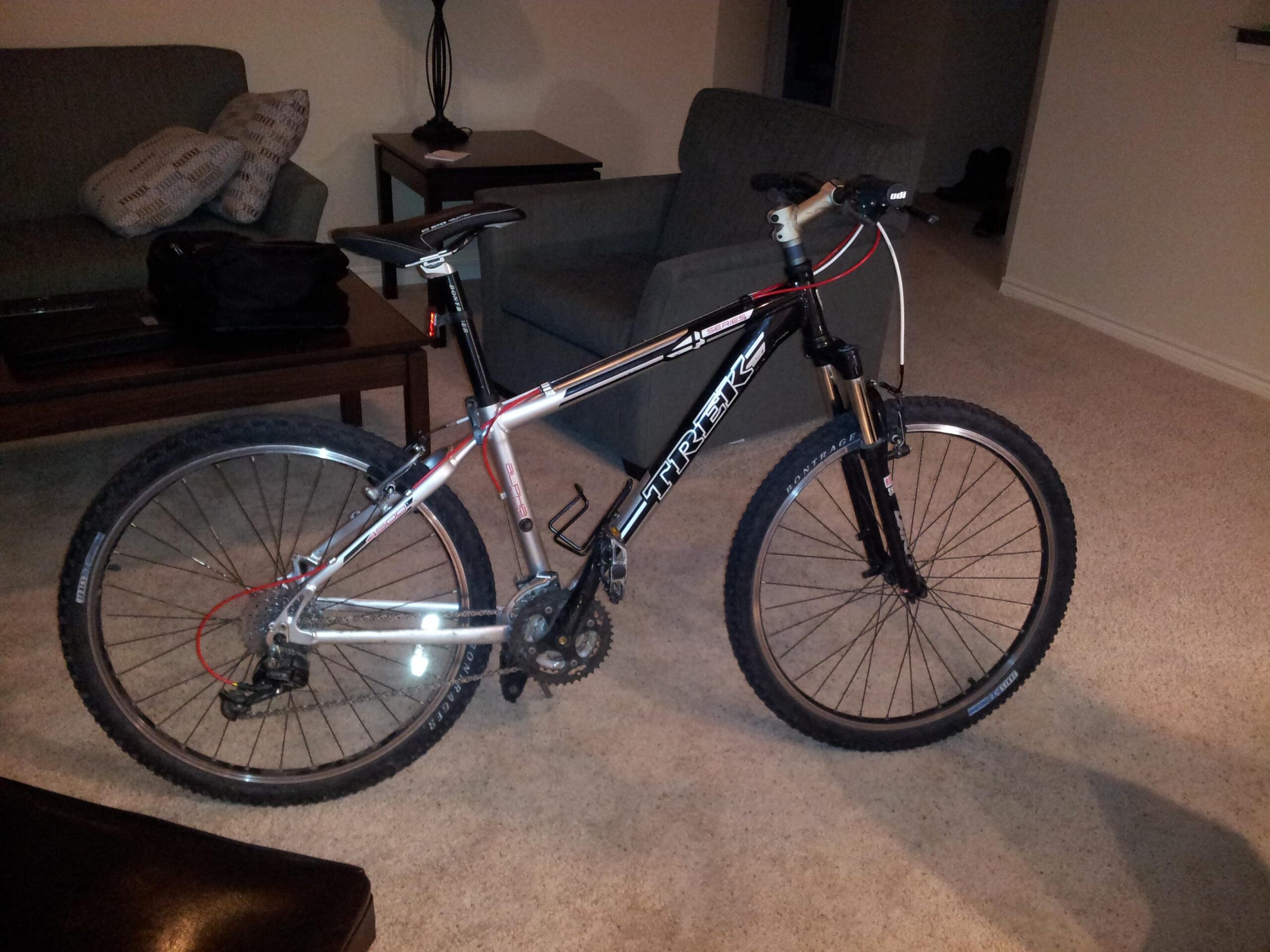 Trek 4500: A black and silver Trek mountain bike is positioned in a living room setting, with carpeted flooring and a couch in the background. A coffee table with a bag and laptop can be seen nearby. The bike features thick tires and a front suspension fork, indicating its suitability for off-road riding.