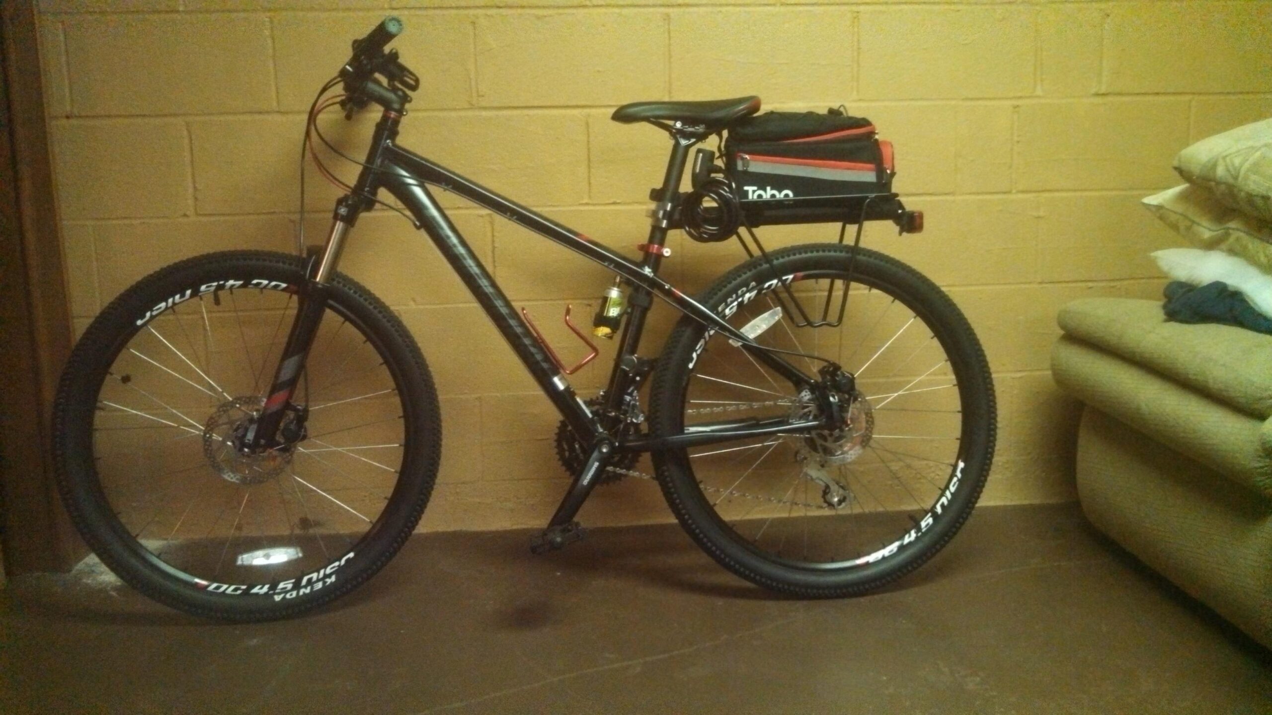 Cannondale Trail 5: A black mountain bike with a red and grey accent is leaning against a beige wall. The bike features a rear rack with a black pouch, a water bottle holder, and thick tires with white lettering. In the background, there is a couch with cushions and a beige wall.