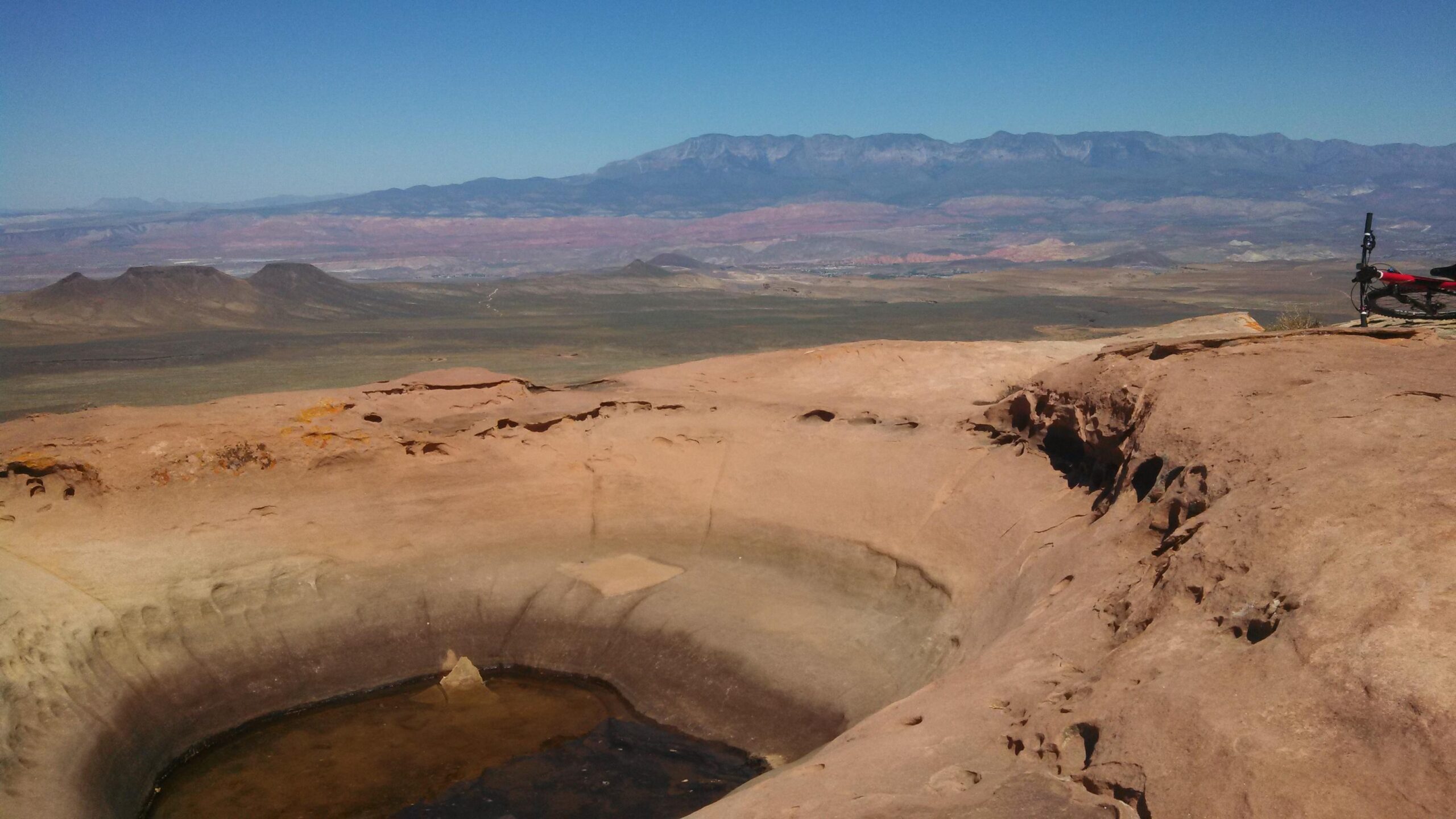 Specialized Stumpjumper FSR Comp Carbon 29: A panoramic view of a rocky landscape featuring a deep circular depression in the foreground and distant mountains under a clear blue sky. The terrain is primarily brown and tan, with some green patches in the expansive valley below. A red mountain bike is visible on the right side of the image.