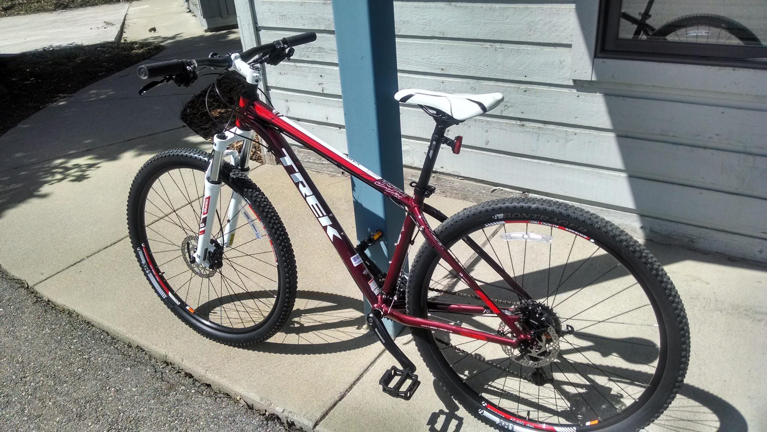 Trek X-Caliber: A red and white mountain bike leaning against a blue post on a sunny day, with a concrete sidewalk and a wooden building in the background. The bike features knobby tires and a modern design.