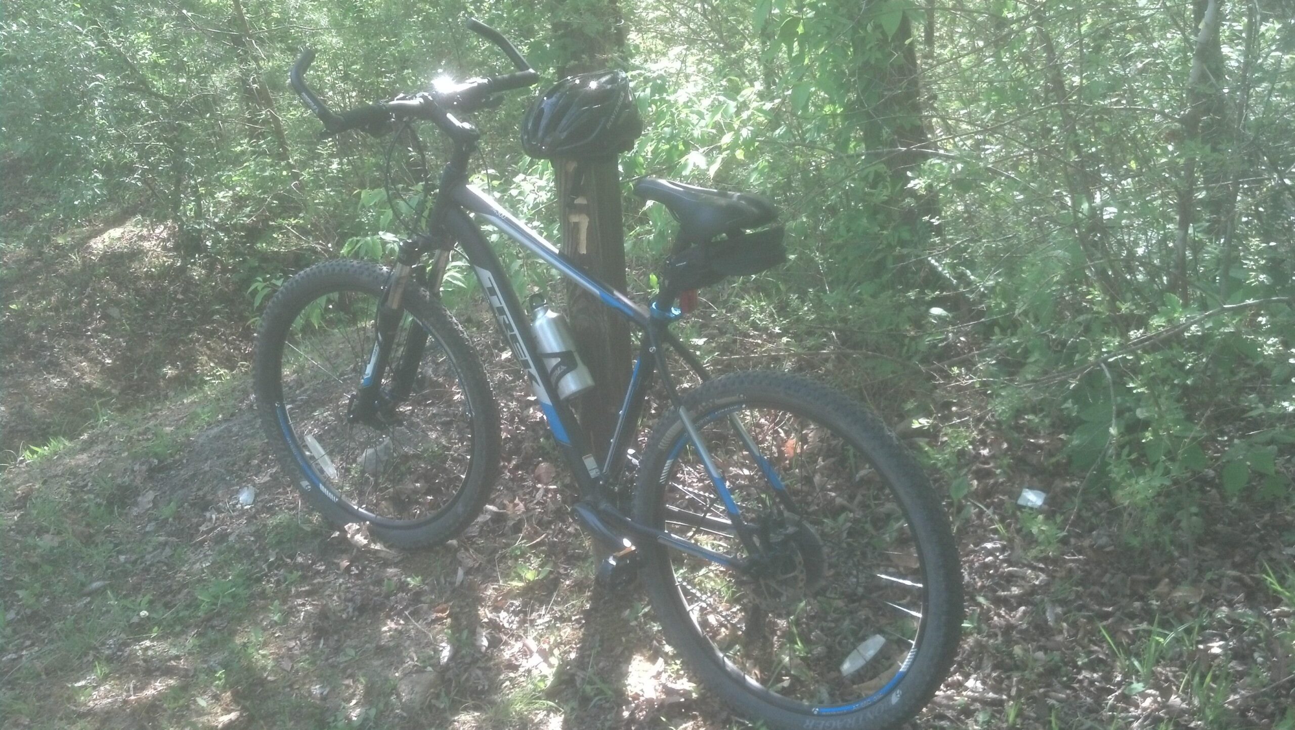Trek 4300: A mountain bike parked next to a wooden post in a forested area, surrounded by greenery and sunlight filtering through the trees. A bicycle helmet is resting on the post, and a water bottle is attached to the bike.
