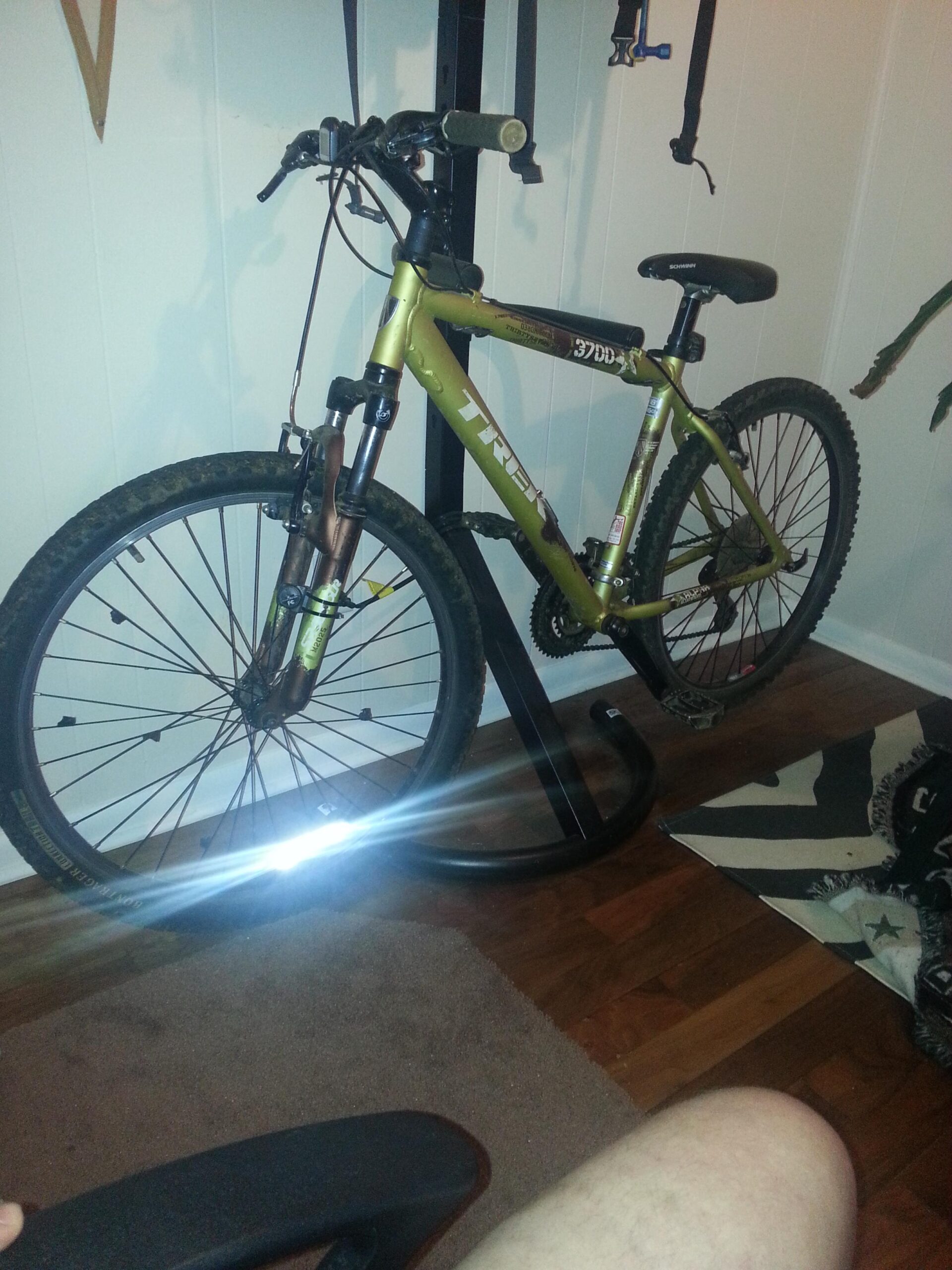 Trek 3700: Alt text: A bicycle with a green frame is leaning against a wall, displaying mud on its tires, indicating recent outdoor use. The floor is wooden, with a brown carpet beneath the bicycle, and parts of a chair and a patterned blanket are visible in the foreground. There is a subtle glare from a light source reflecting off the bicycle's wheel.