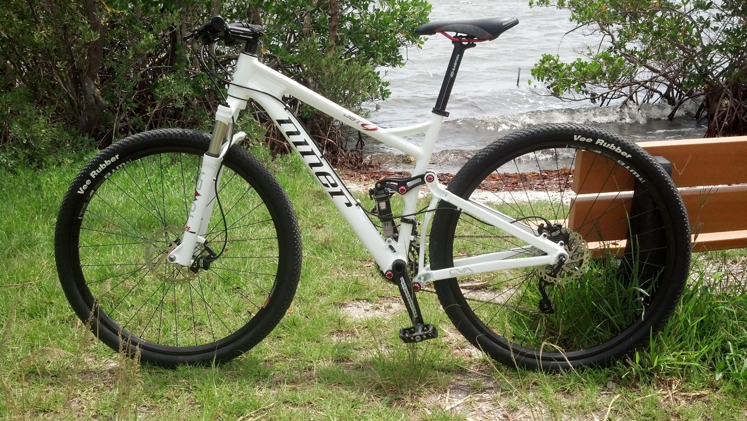 Niner Jet 9: A white mountain bike parked on green grass, with a wooden bench and a body of water in the background. The bike features a suspension system, disc brakes, and black tires with "Vee Rubber" branding. Lush vegetation surrounds the scene, creating a natural outdoor setting.