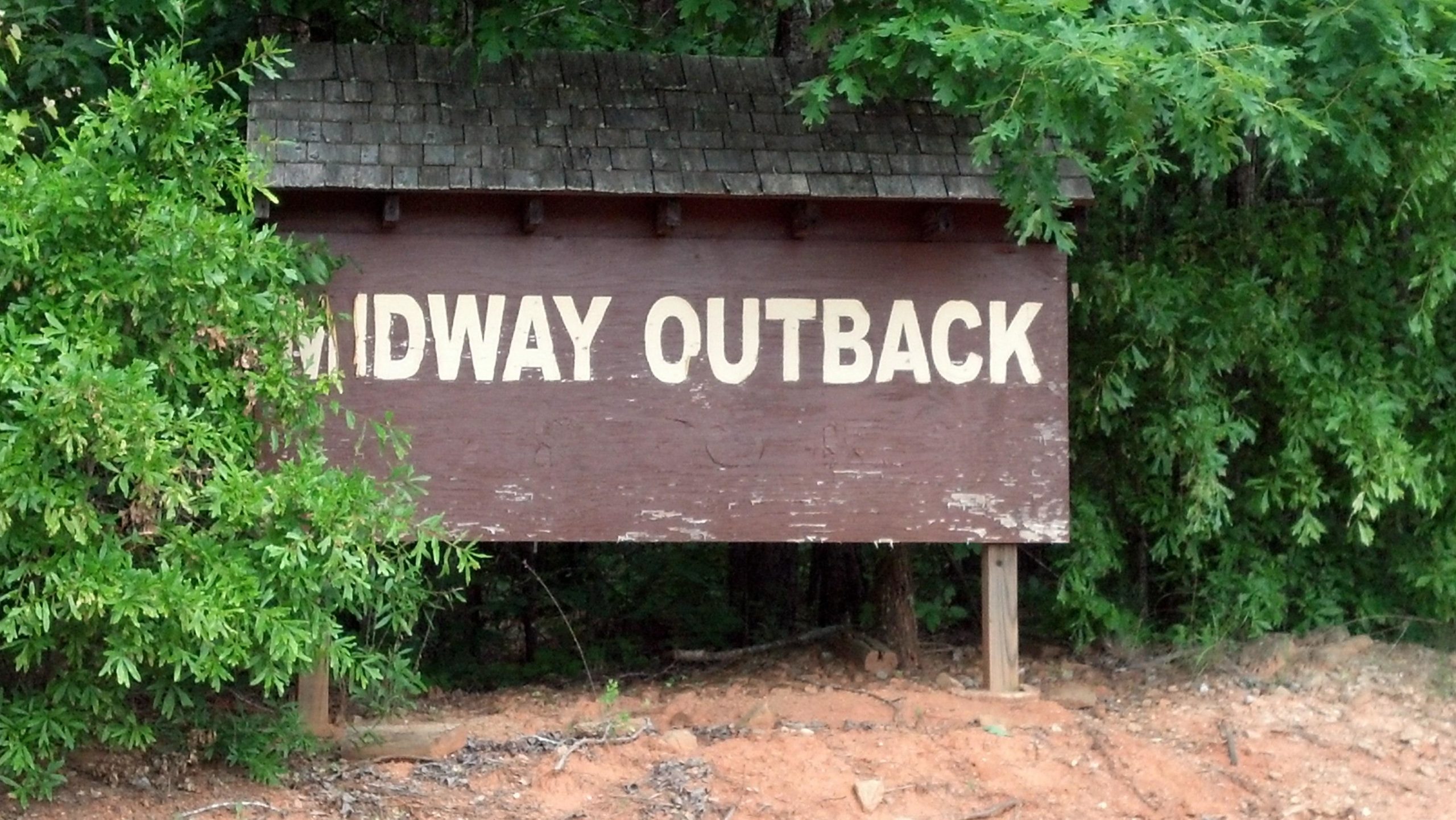 Wooden sign reading "MIDWAY OUTBACK," partially obscured by green foliage. The sign is set in a natural outdoor environment. Midway Outback mountain bike trail.