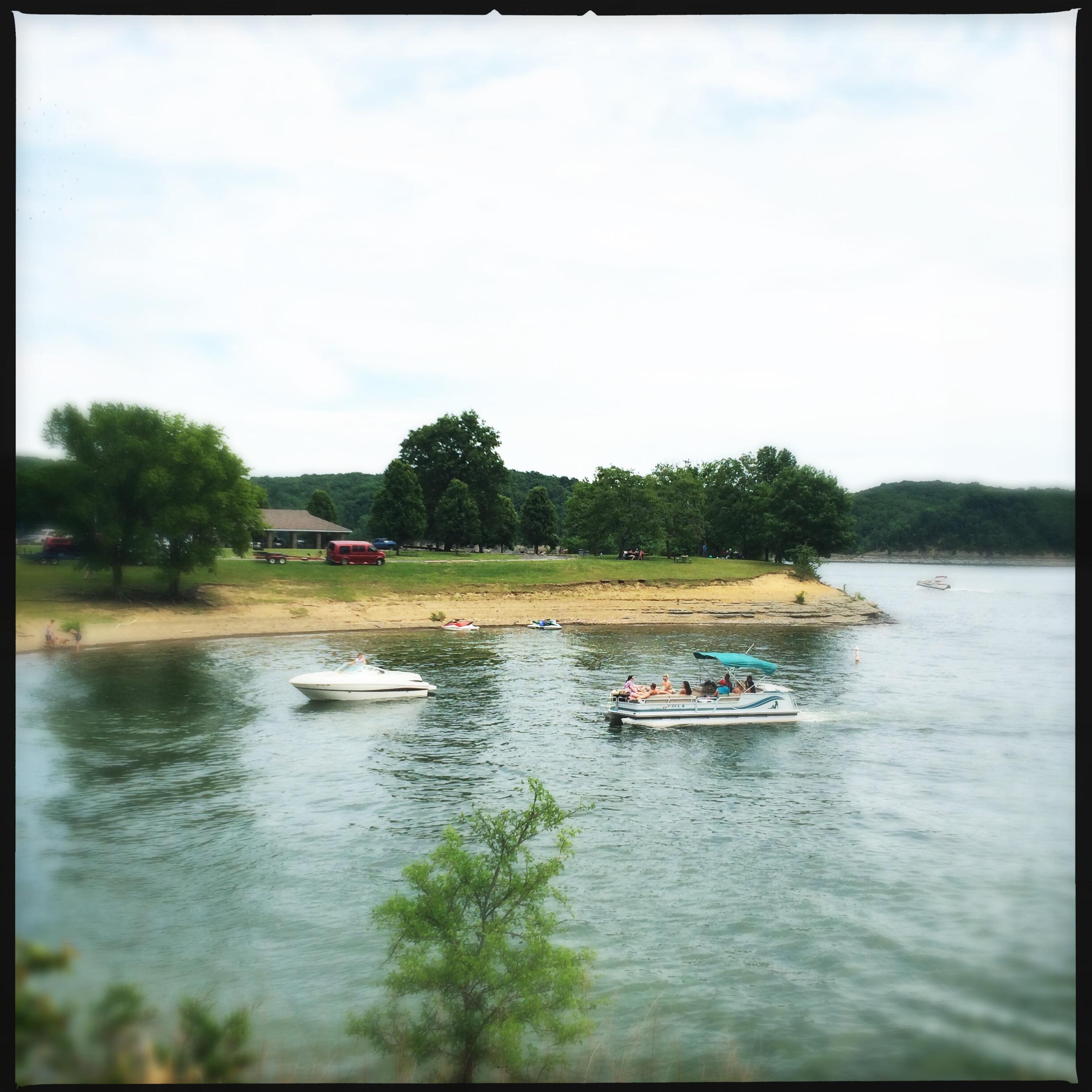 A scenic view of a lake with two boats on the water. In the background, there is a sandy beach with people relaxing and enjoying the outdoors, surrounded by green trees and hills. A grassy area with a parking lot and a building can be seen on the shore. The sky is partly cloudy, creating a tranquil atmosphere. Green River mountain bike trail.