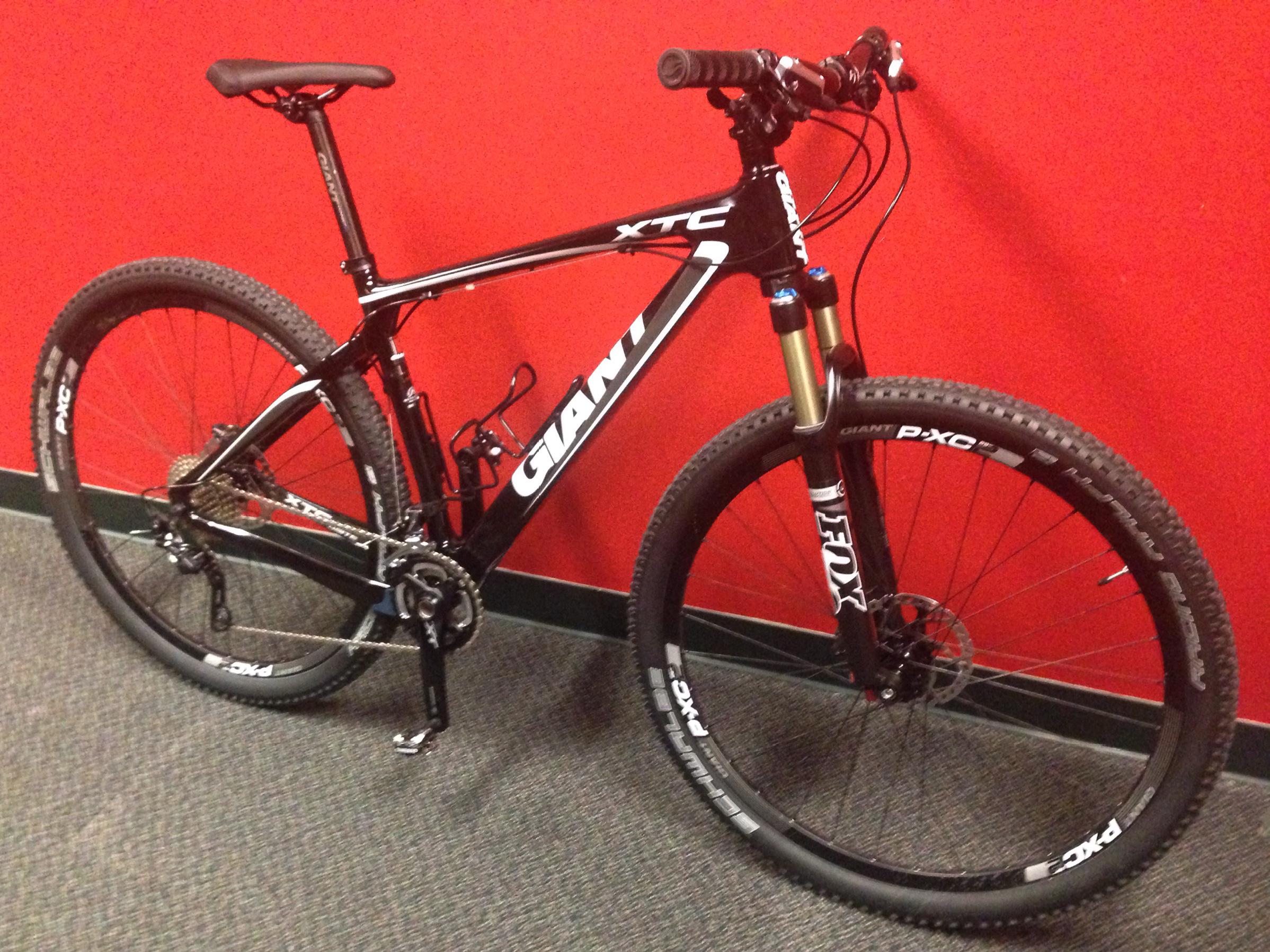 Giant XTC Composite 29er 1: A black Giant XTC mountain bike positioned against a red wall. The bike features thick tires and front suspension components, showcasing a modern design suitable for off-road cycling. The pedals and gears are visible, highlighting its geared system.