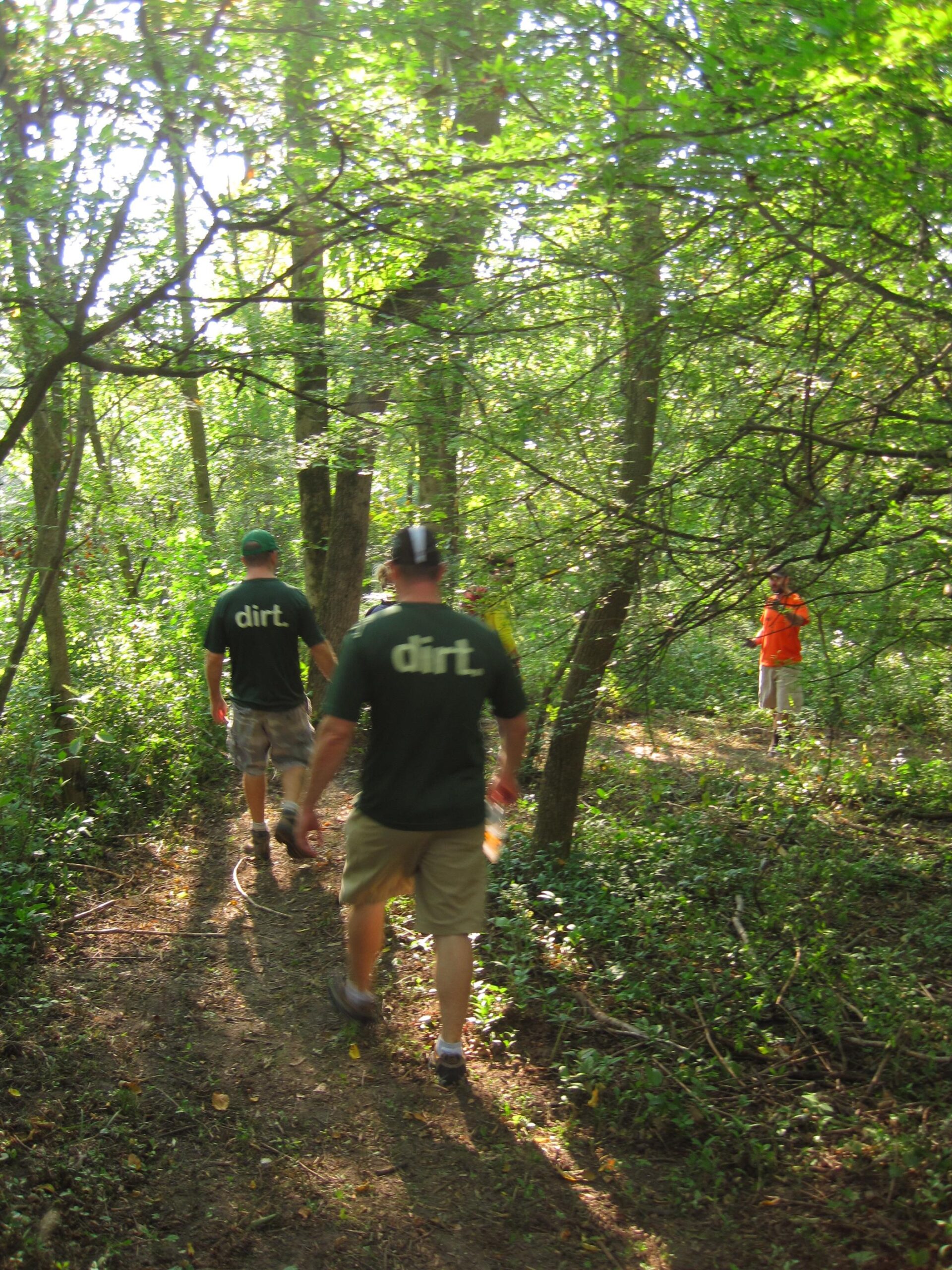 A group of people walking along a dirt path in a lush, green forest. Two individuals in dark shirts with the word "dirt" printed on them are walking towards the camera, while a person in an orange shirt can be seen in the background. Sunlight filters through the trees, creating a serene outdoor atmosphere.