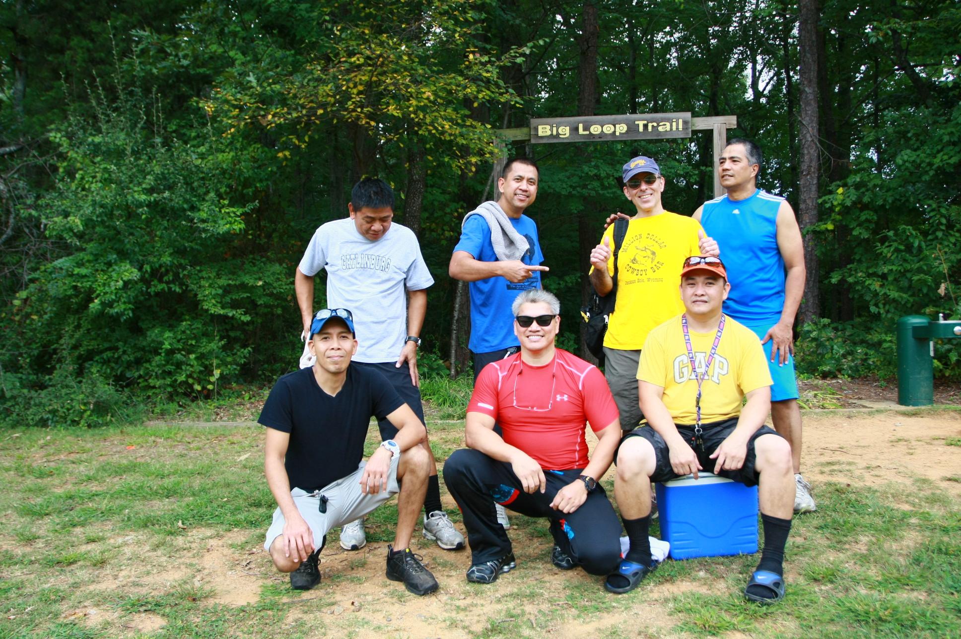 A group of seven men posing together at the Big Loop Trail sign in a wooded area. They display a variety of clothing suitable for outdoor activities, with smiles and playful gestures indicating a friendly atmosphere. The background features greenery and trees typical of a natural park setting. Big Loop mountain bike trail.