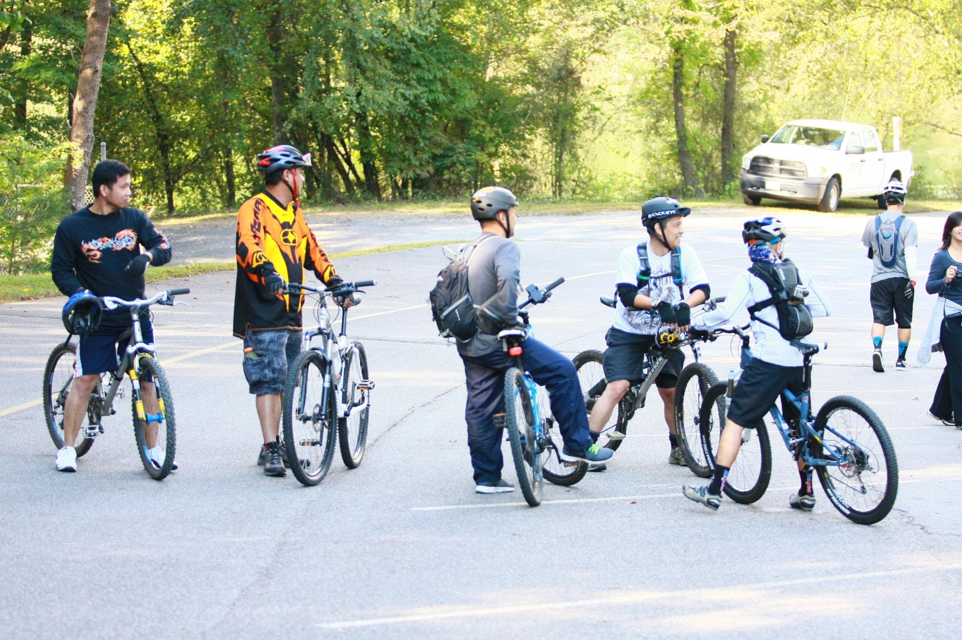A group of six individuals, wearing cycling gear, socialize in a parking area surrounded by trees. They are standing near their mountain bikes, with some engaged in conversation and others preparing to ride. The scene is set in a bright, outdoor environment, indicating a pleasant day for biking. Overmountain Victory Trail mountain bike trail.