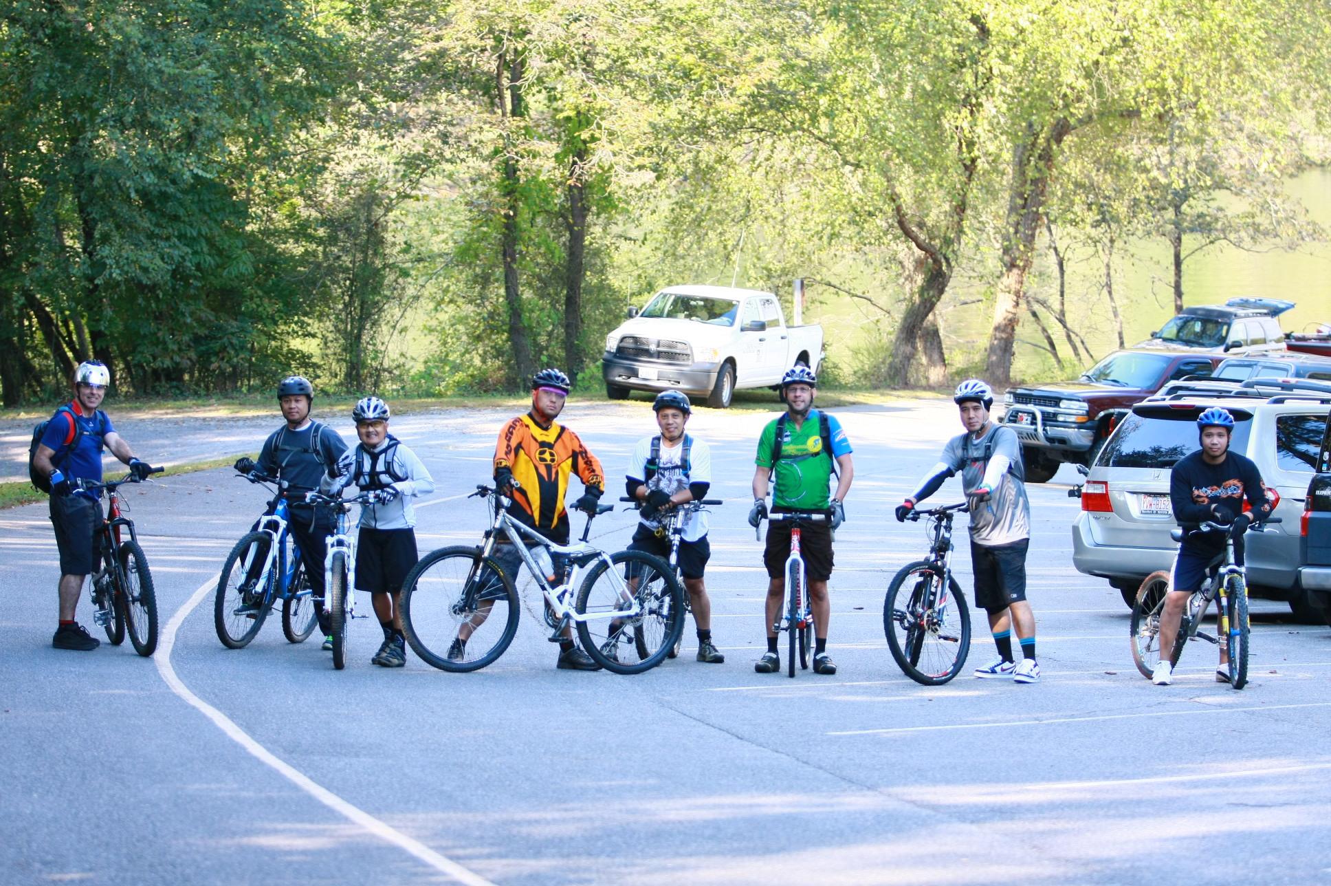 A group of seven cyclists, wearing helmets and riding gear, pause on a paved path in a shaded parking area. They are standing next to their mountain bikes, with trees and vehicles visible in the background. The scene captures a moment of camaraderie among the riders. Overmountain Victory Trail mountain bike trail.