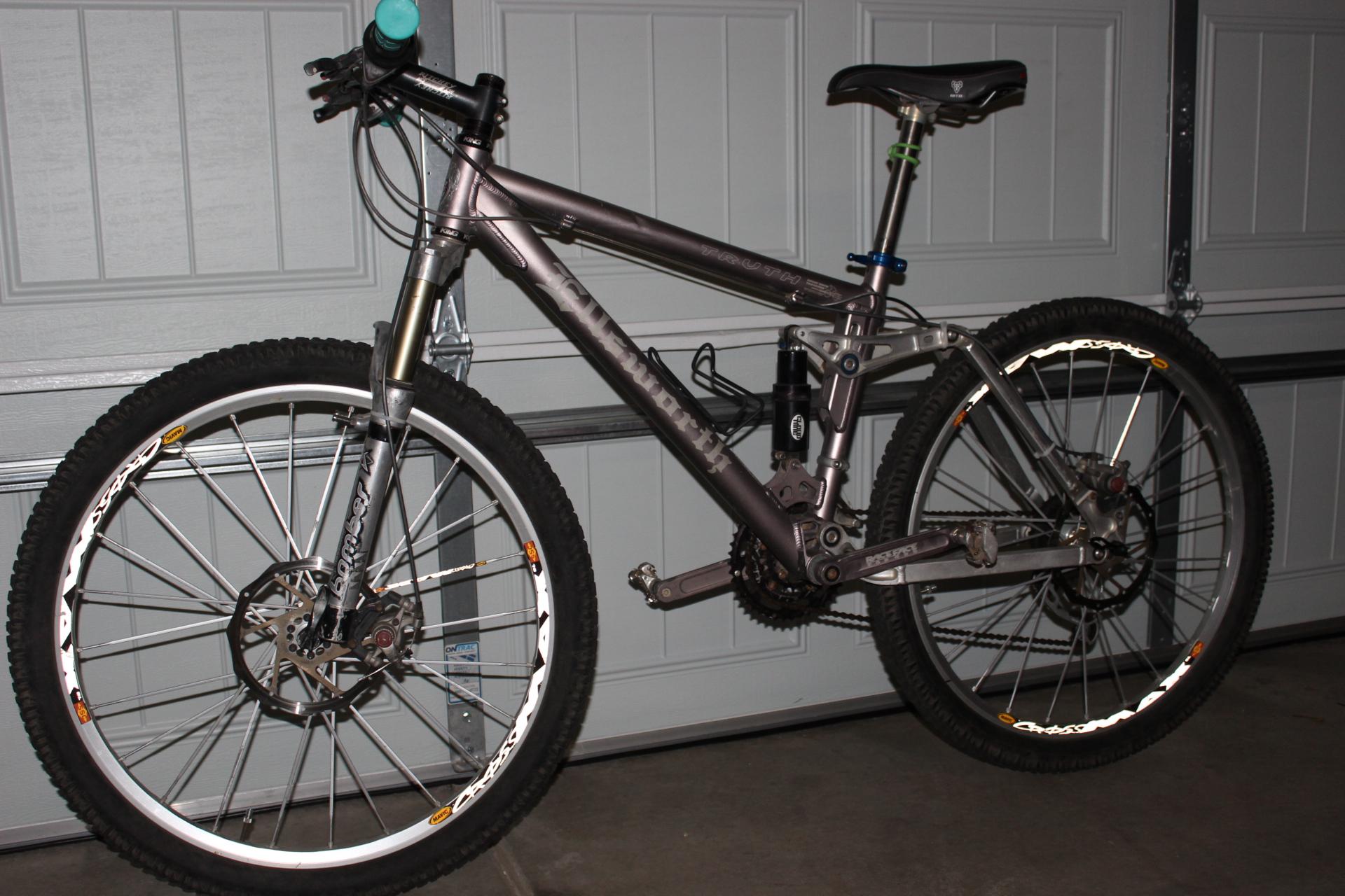 Ellsworth Truth: A parked mountain bike with a silver frame and distinct shock absorber, set against a garage door background. The bike features thick tires with visible tread and disc brakes. The handlebars are equipped with grips and controls, and the seat is adjustable.