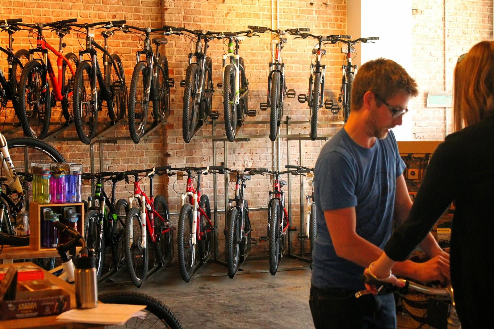 An interior view of a bike shop featuring a variety of bicycles displayed on racks against a brick wall. Two individuals are engaged in a conversation, with one person handling a bicycle while the other appears to be assisting. Various bicycle accessories and drinks are visible on a counter in the foreground. The ambiance is warm and inviting, with natural light highlighting the setting.