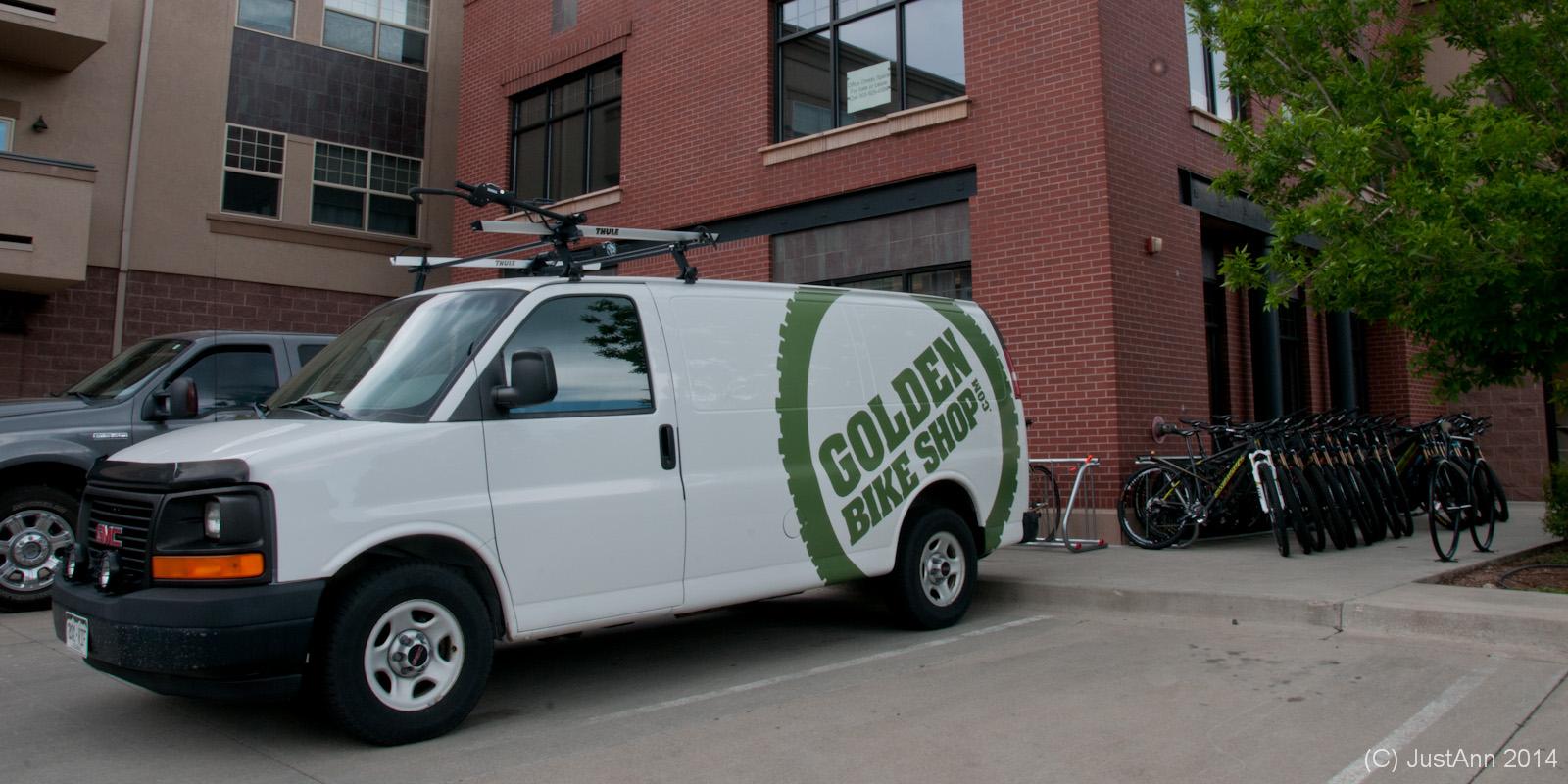 A white van with "Golden Bike Shop" branding parked in front of a brick building, with bicycles lined up in a rack beside it. The van features a bike rack on the roof and the building's windows are visible behind it.
