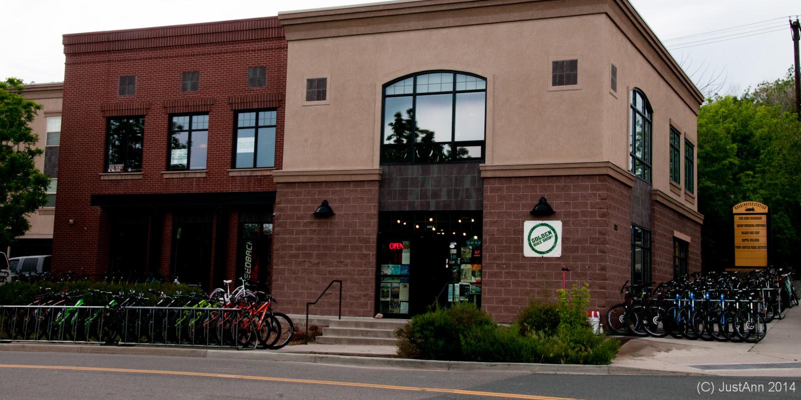A two-story building with a combination of brick and tan stucco exterior. The ground level has large windows and an "OPEN" sign visible. In front of the building, there is a wide bike rack filled with a variety of colorful bicycles. Lush green plants and trees are visible nearby, along with a sign on the right indicating local businesses.