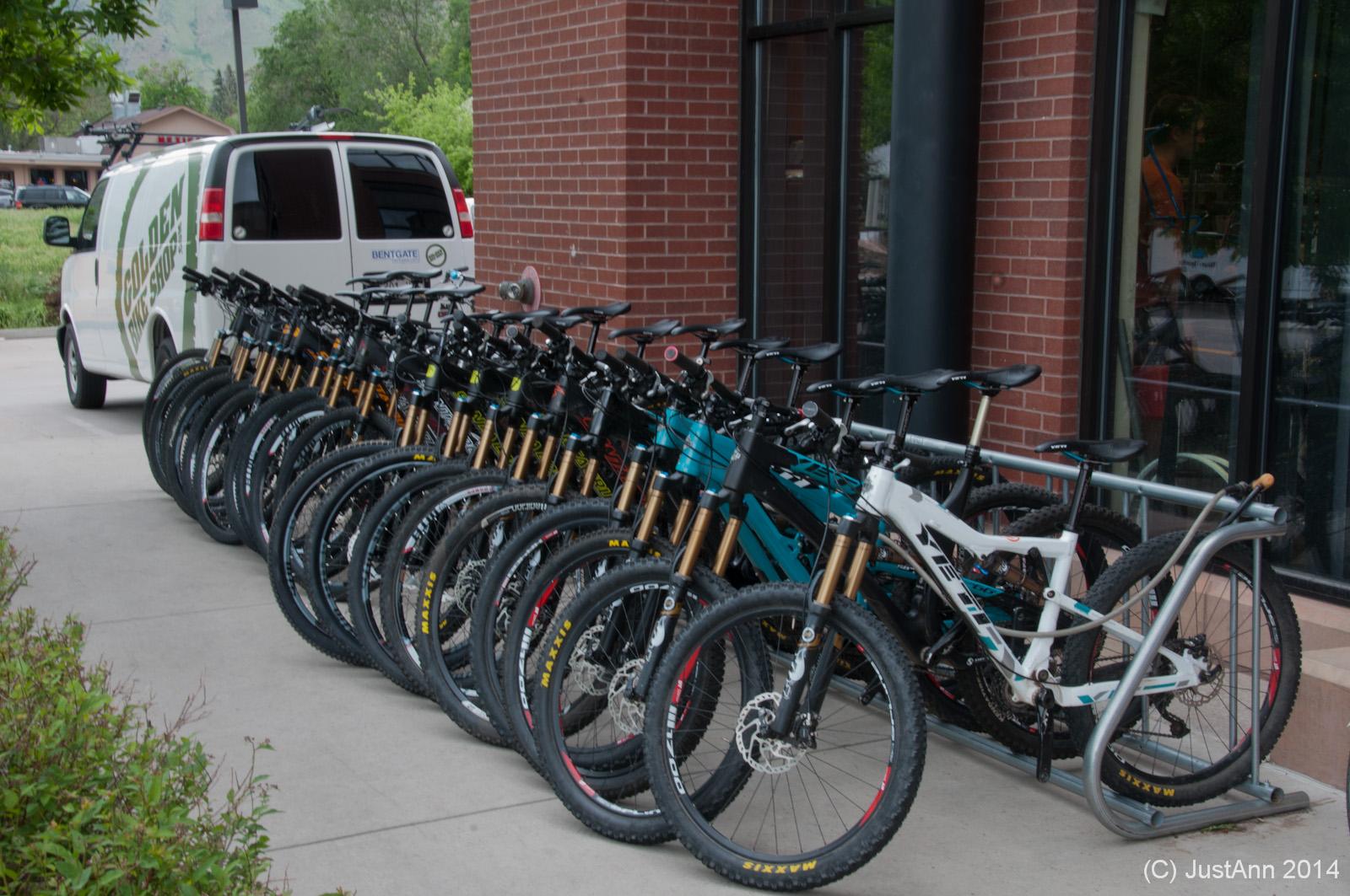 A row of mountain bikes in various colors is lined up against a bike rack outside a brick building, with a white van featuring a logo parked nearby. The area is surrounded by greenery, indicating an outdoor setting.