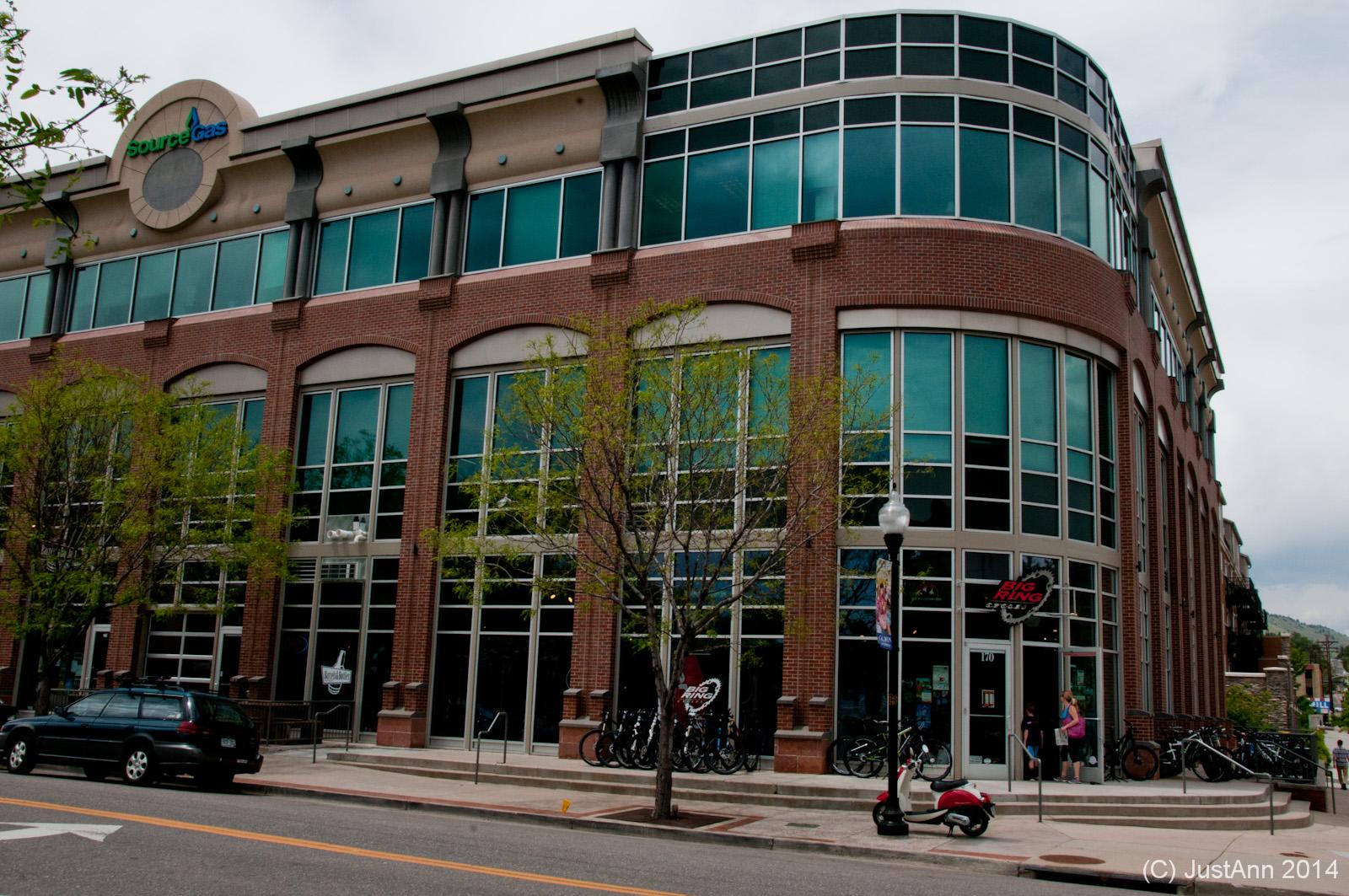 A modern brick building with large glass windows, featuring the SourceGas logo at the top. The entrance has a sign for "Big Ring" and is surrounded by trees and parked bicycles. A few people are seen near the entrance, with a parked car and scooter on the street. The sky is overcast, adding a soft gray tone to the scene.
