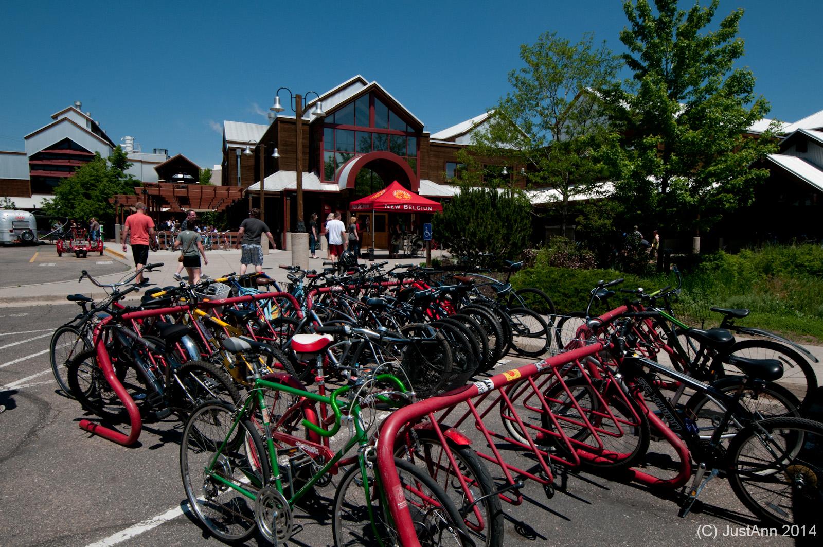 A bustling scene outside a building featuring a large, red canopy with the logo "New Belgium." In the foreground, multiple bicycles are parked in a designated area with a unique bike rack, while several people are walking toward the entrance. The sky is clear and blue, indicating a sunny day.