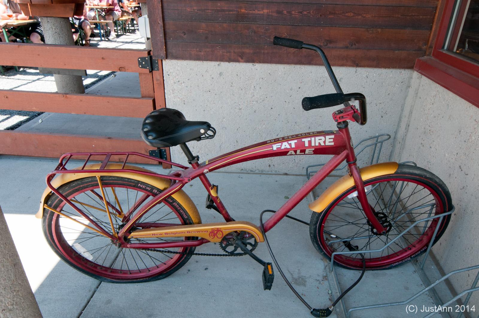 A vintage-style bicycle with a bright red frame and yellow accents, featuring the text "Fat Tire Ale," resting against a wall near a wooden gate. The bike has a classic design with a comfortable seat and reflects a laid-back, outdoor setting. In the background, a blurred view of people at tables can be seen.