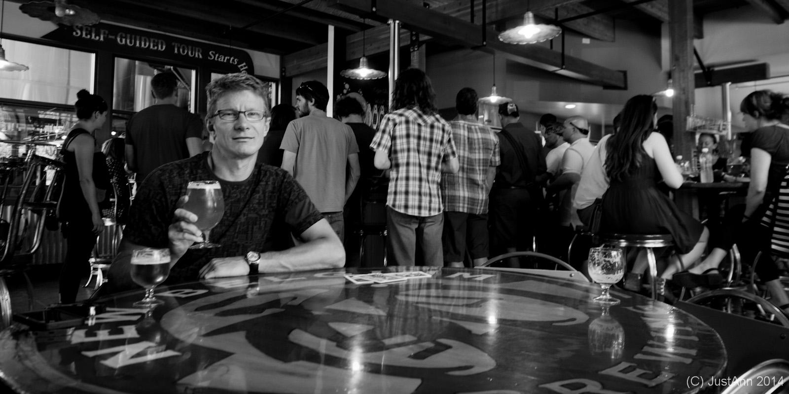 A man sitting at a table in a brewery or pub, holding a glass of beer, with a bustling crowd in the background. The scene is in black and white, showcasing the lively atmosphere of the establishment, with people engaged in conversation and enjoying their drinks. The table has a logo visible, adding to the setting's character.