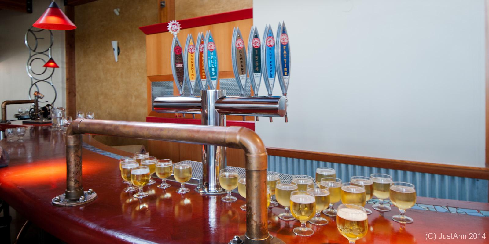 Interior of a bar featuring a polished wooden counter with a row of beer tap handles above. Several glasses of pale beer are arranged on the bar, showcasing clear liquid and foamy tops. The ambiance includes modern lighting with red pendant lamps and a stylish wall design.