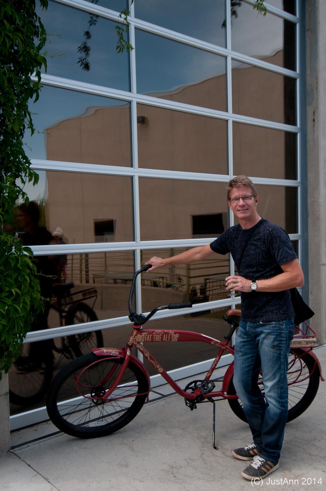 A man standing beside a red bicycle with the words "Fat Tire Ale" on it, smiling at the camera. He is holding a drink in one hand and has a casual outfit consisting of a black shirt and blue jeans. The reflection of a building and greenery can be seen in the large glass windows behind him.