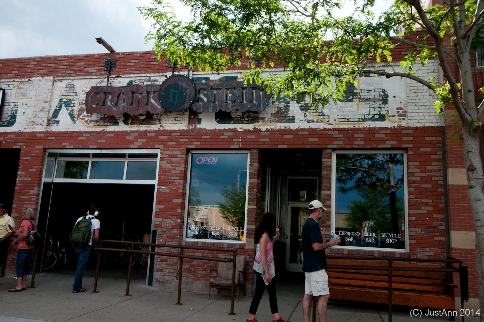 Alt text: Exterior view of a brick storefront featuring a sign that reads "Craft," with an open sign in the window. In front, a few people are standing, including a woman and a man with a backpack. There is a bench and some greenery visible nearby.