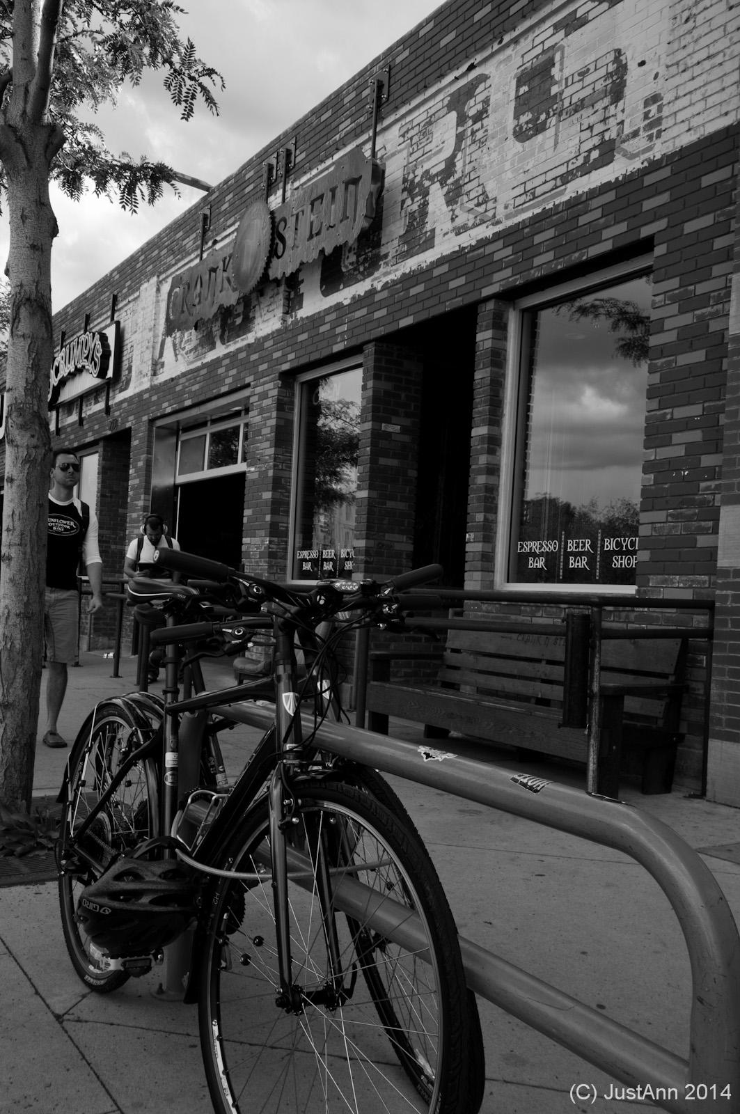 Bicycles are parked at a bike rack in front of a brick-building café with an old sign partially visible. Two people walk by on the sidewalk, and a bench is placed next to the entrance, which has large windows displaying café-related signage. The image is in black and white.
