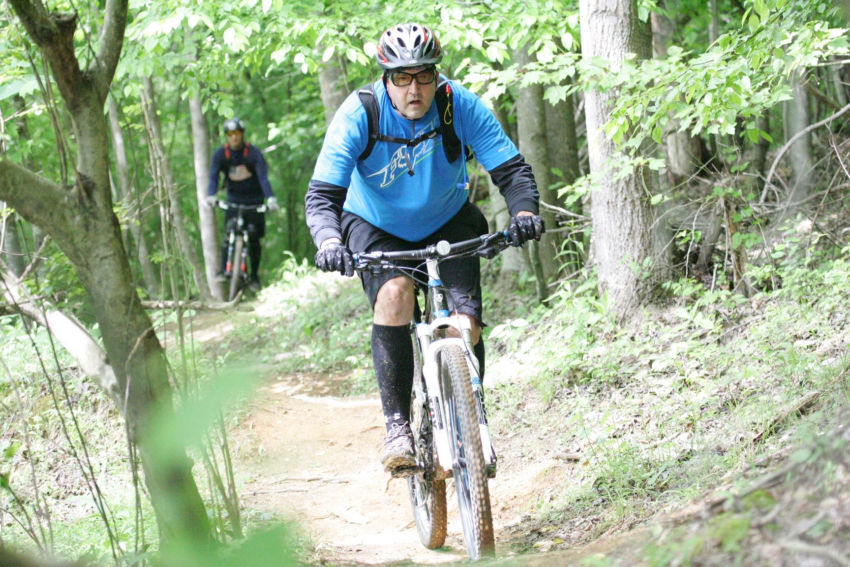 A mountain biker wearing a blue shirt and a helmet rides along a forest trail, surrounded by green foliage. A second cyclist can be seen in the background. Warrior Creek mountain bike trail.