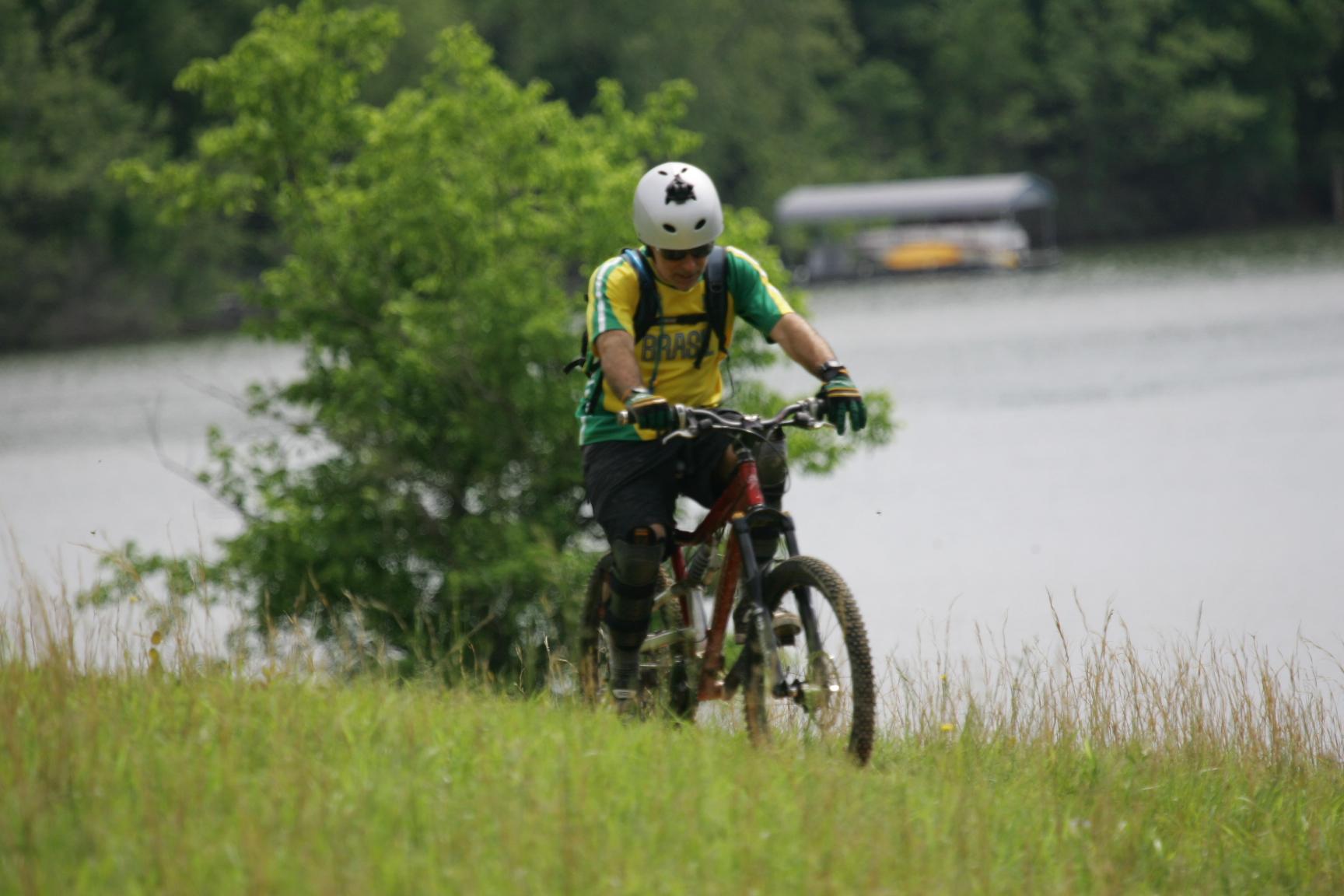 A mountain biker wearing a helmet and protective gear rides down a grassy slope near a lake, surrounded by trees. The biker is dressed in a yellow and green jersey and appears focused as he navigates the terrain. Warrior Creek mountain bike trail.