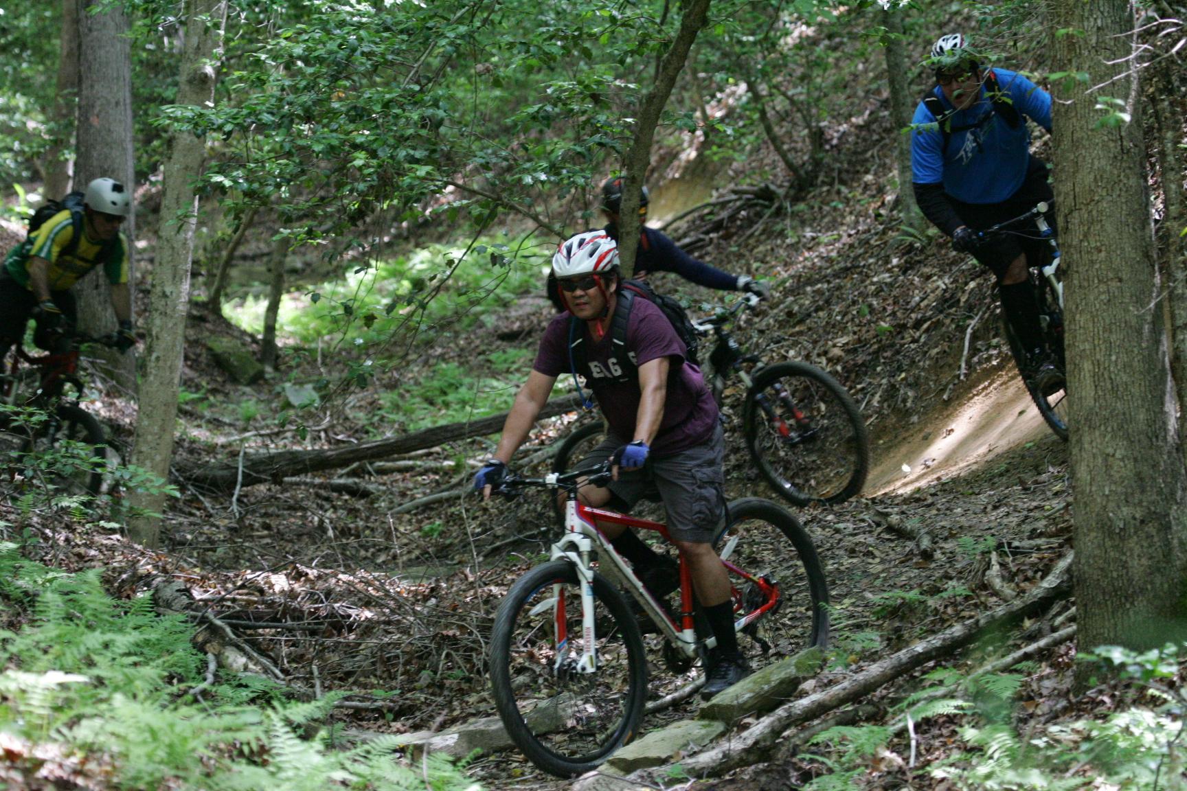 A group of mountain bikers navigating a rocky trail through a wooded area, surrounded by greenery and trees. The riders are wearing helmets and protective gear, showcasing an active outdoor sport in a nature setting. Warrior Creek mountain bike trail.
